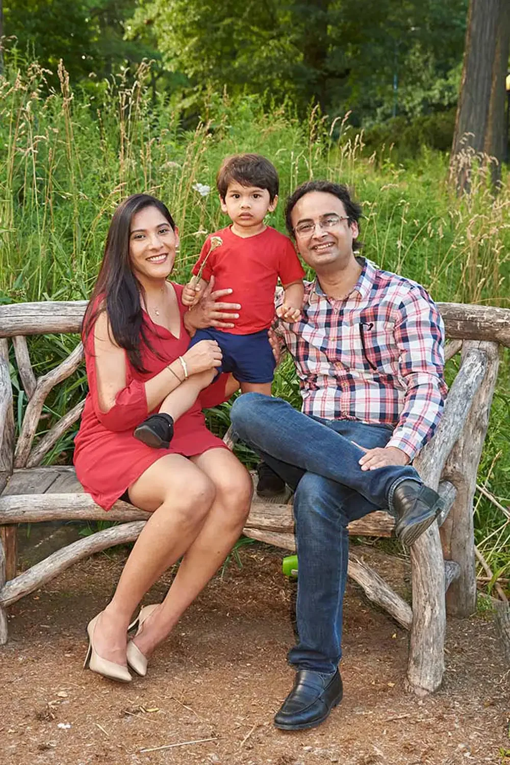 A family of three sitting on a rustic wooden bench outdoors surrounded by green grass and trees. The woman is wearing a red dress and beige heels, holding her son on her lap. The young boy is wearing a red t-shirt and blue shorts.