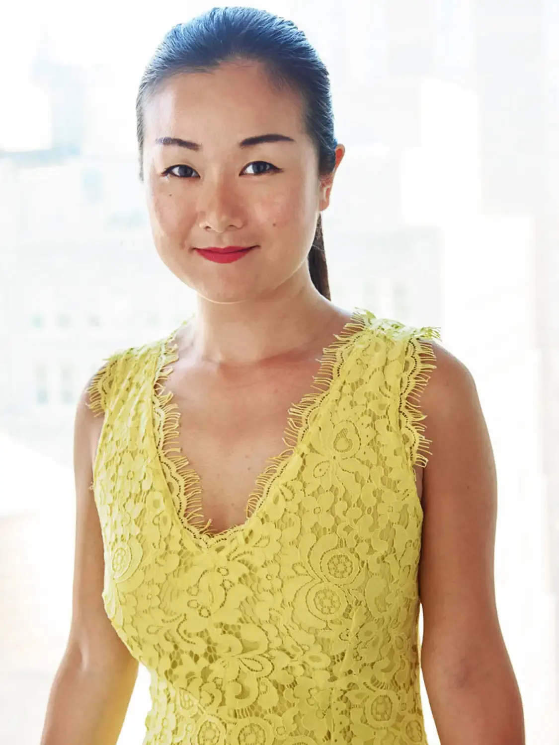 A headshot of a. woman with dark hair pulled back, wearing a yellow lace sleeveless dress, smiling in front of a bright, sunny window.