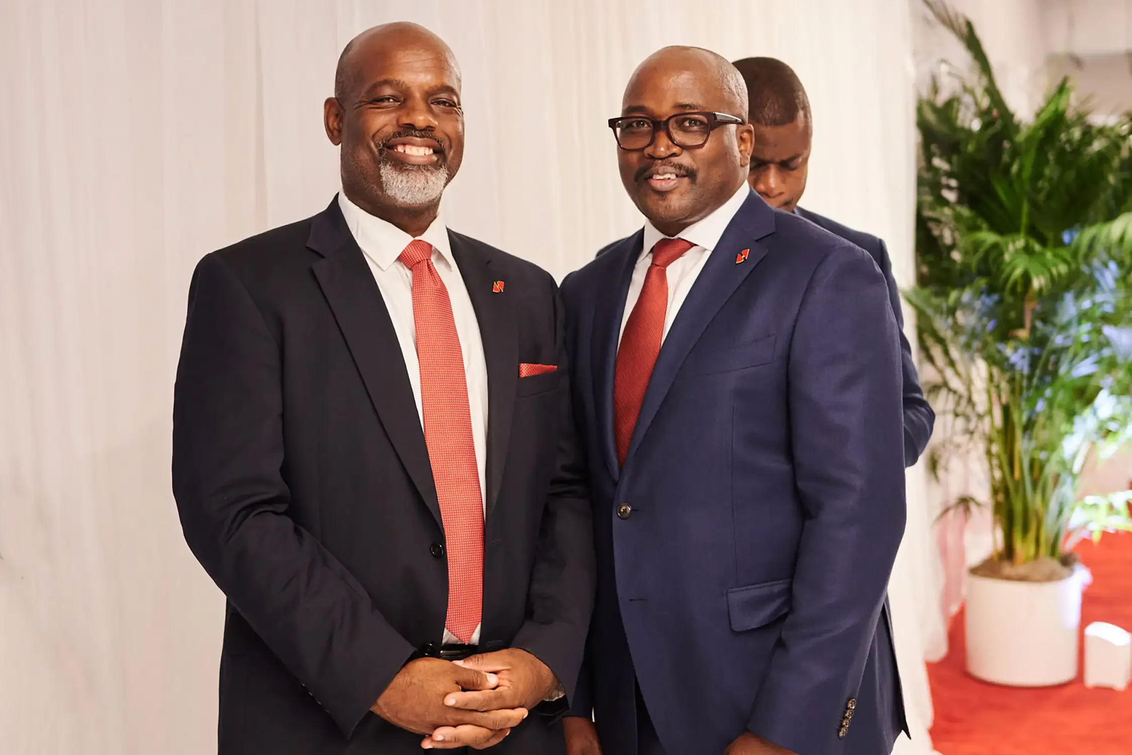 Two men in formal suits with red ties and lapel pins smiling and standing close together in a bright room, with a third man blurred in the background, at a corporate event nyc.
