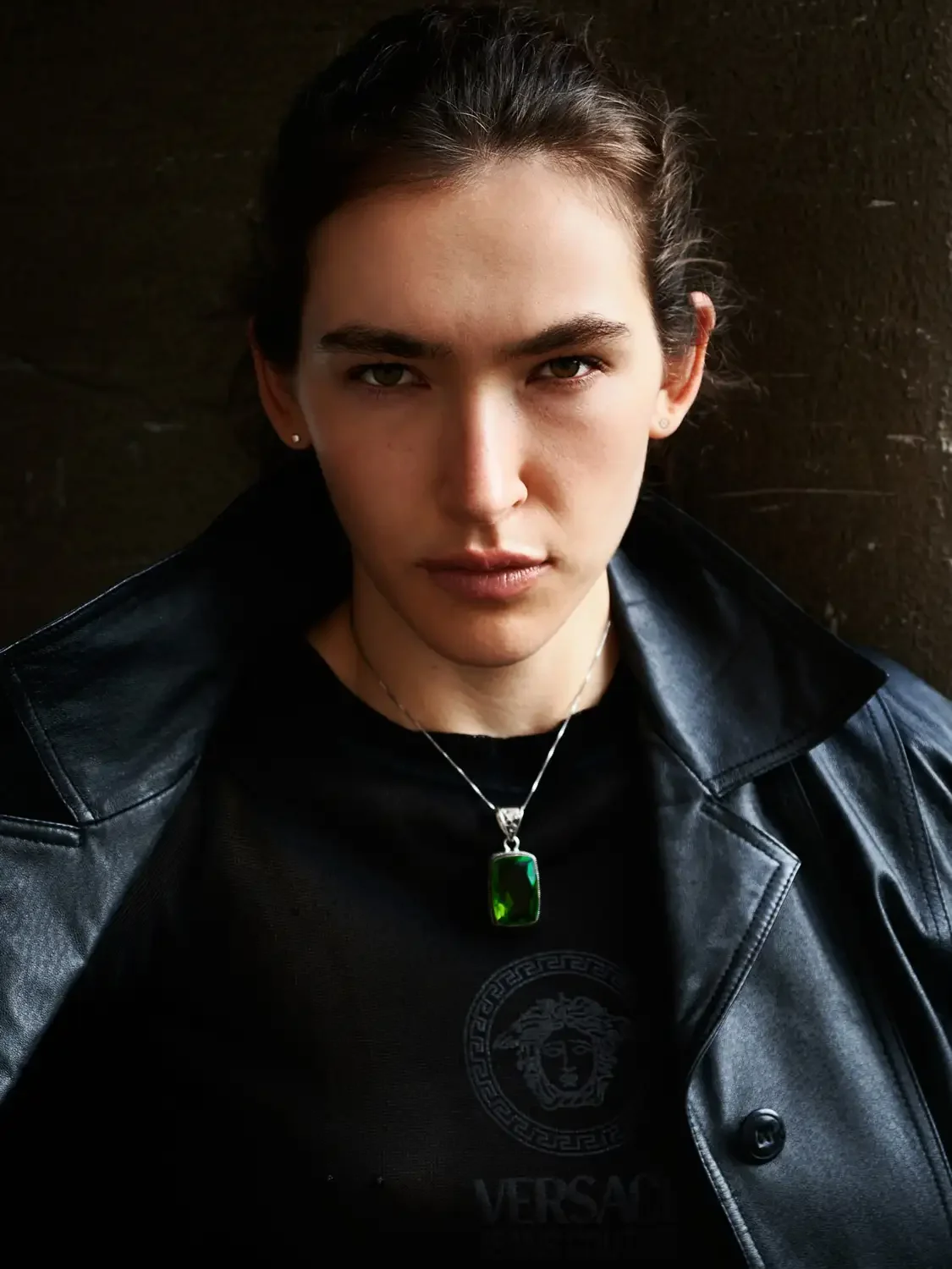 A young woman with dark hair pulled back, wearing a black leather jacket, a black Versace t-shirt, and a necklace with a large green gemstone pendant, looking directly at the camera with a serious expression.