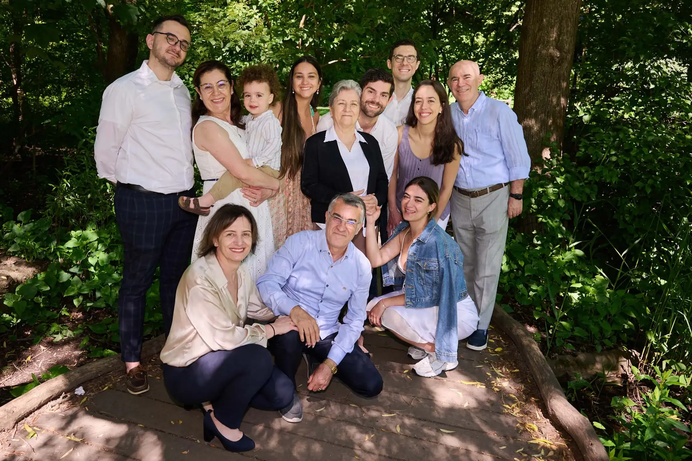 A group of sixteen people, including children, adults, and seniors, gathered in a lush, green outdoor setting for a family photo. They are smiling and standing close together in Central Park New York