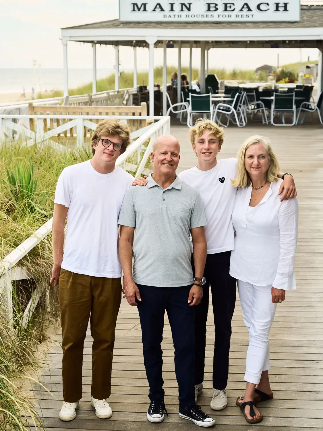Photograph of a family of four standing on a wooden boardwalk in East Hampton, with a sign above them reading "Main Beach Bath Houses for Rent." The family is smiling and dressed casually in white and light-colored clothes.