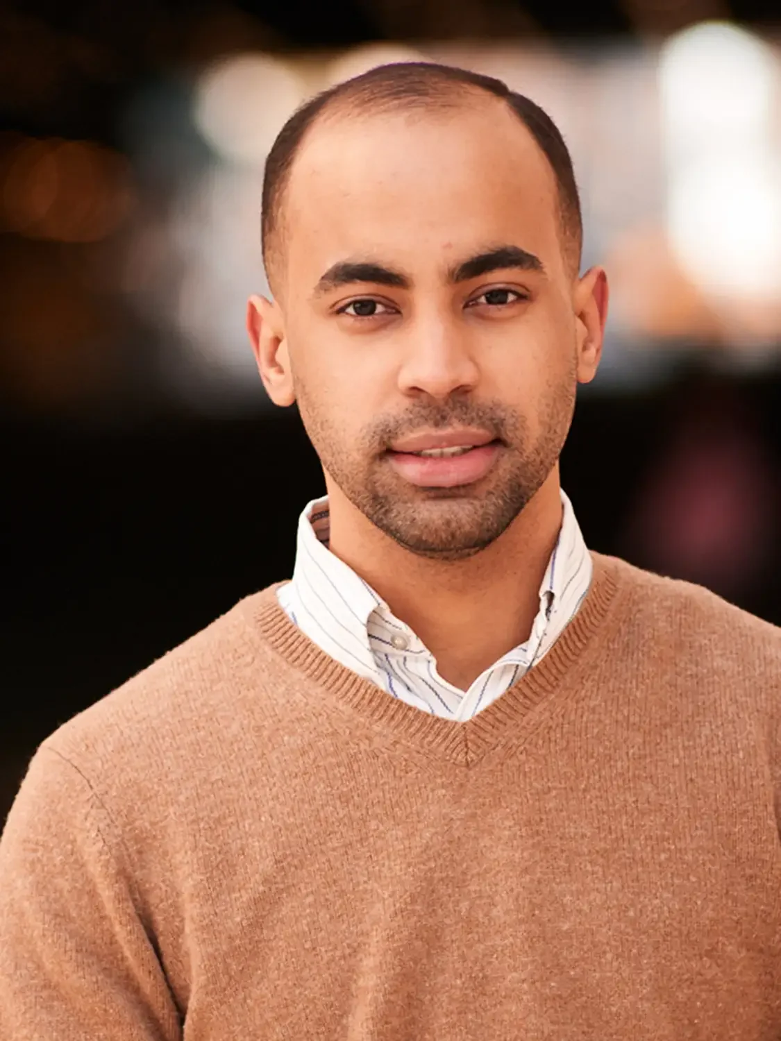 Professional headshot of a young man with short dark hair, light brown skin, wearing a beige sweater over a white collared shirt, standing indoors with a blurred background.