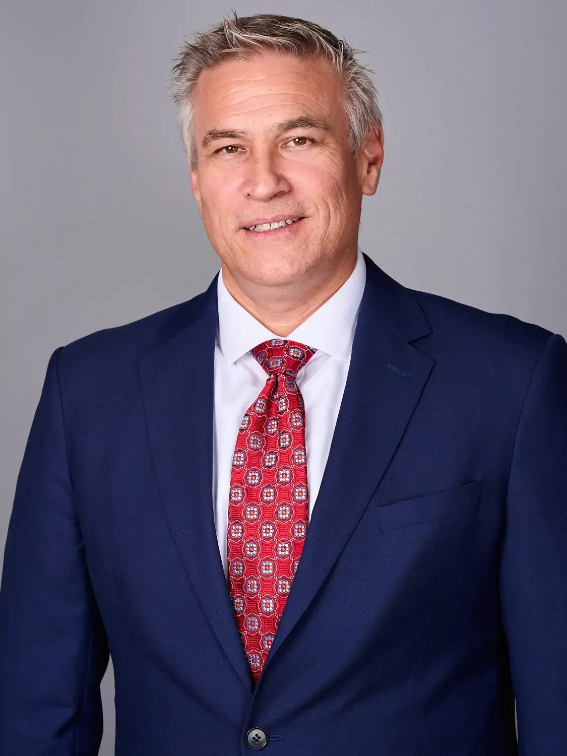 Headshot of a middle-aged executive  in a formal navy blue suit with a white shirt and a red patterned tie, photographed in a professional photo studio.