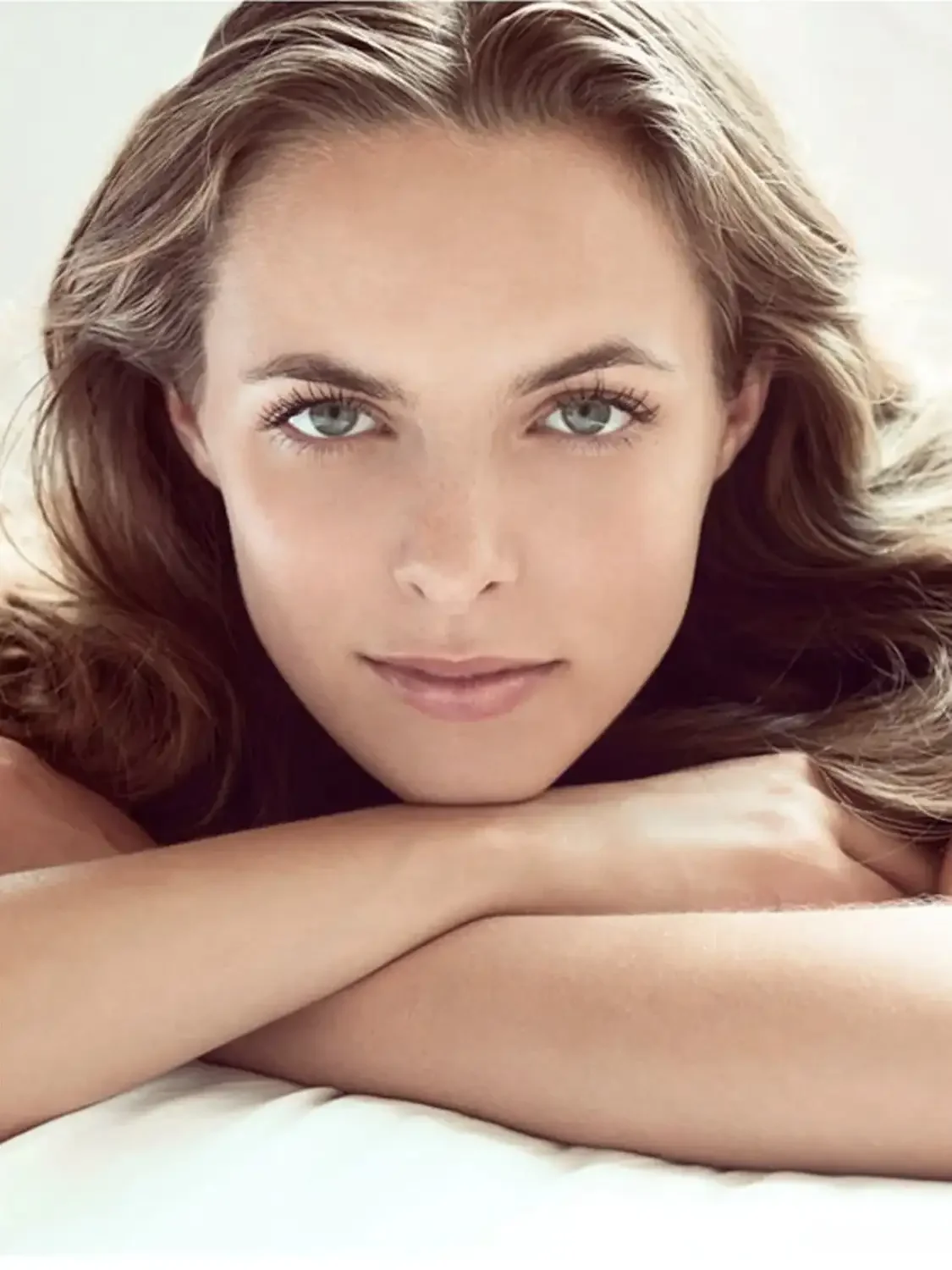 Beauty shot of a young woman with fair skin and light brown hair resting her head on her crossed arms, looking directly at the camera for a cosmetic advertising.