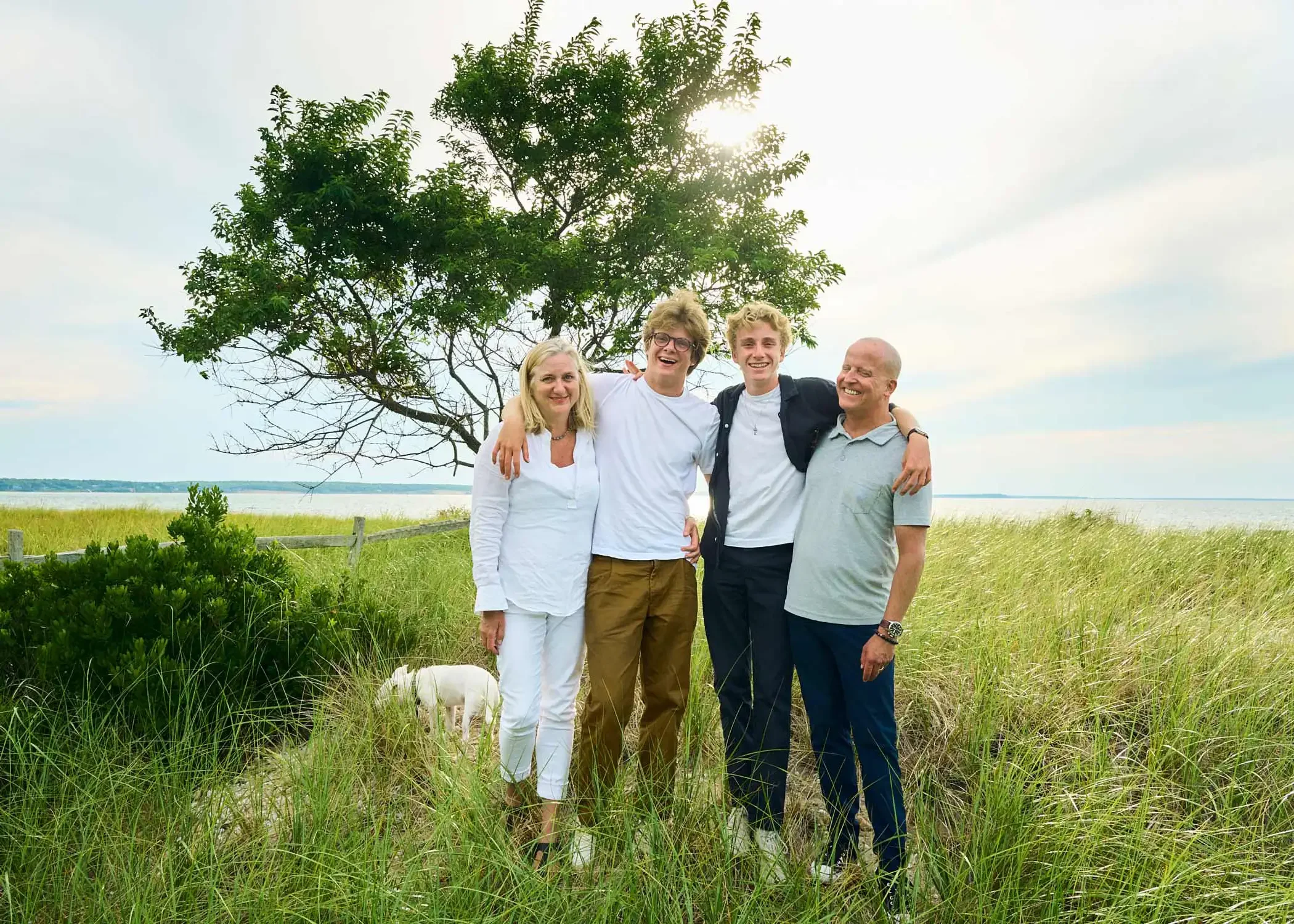 Photo of four people standing together outdoors near a body of water with trees and grass in the background. One person is holding a small white dog.