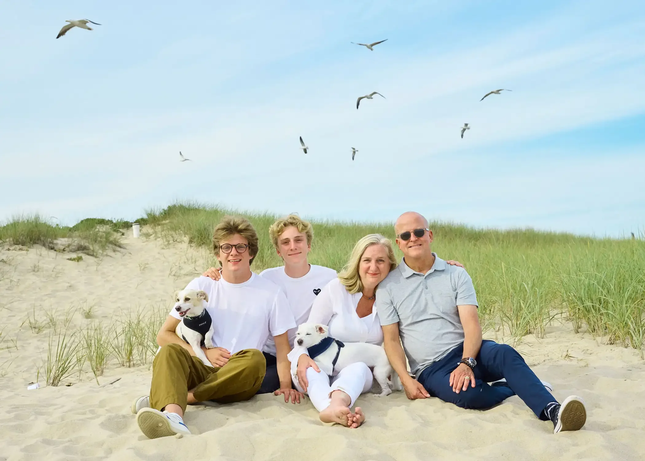 A group of five people photographed sitting on the sand at the beach with grassy dunes in the background and seagulls flying in the sky above.