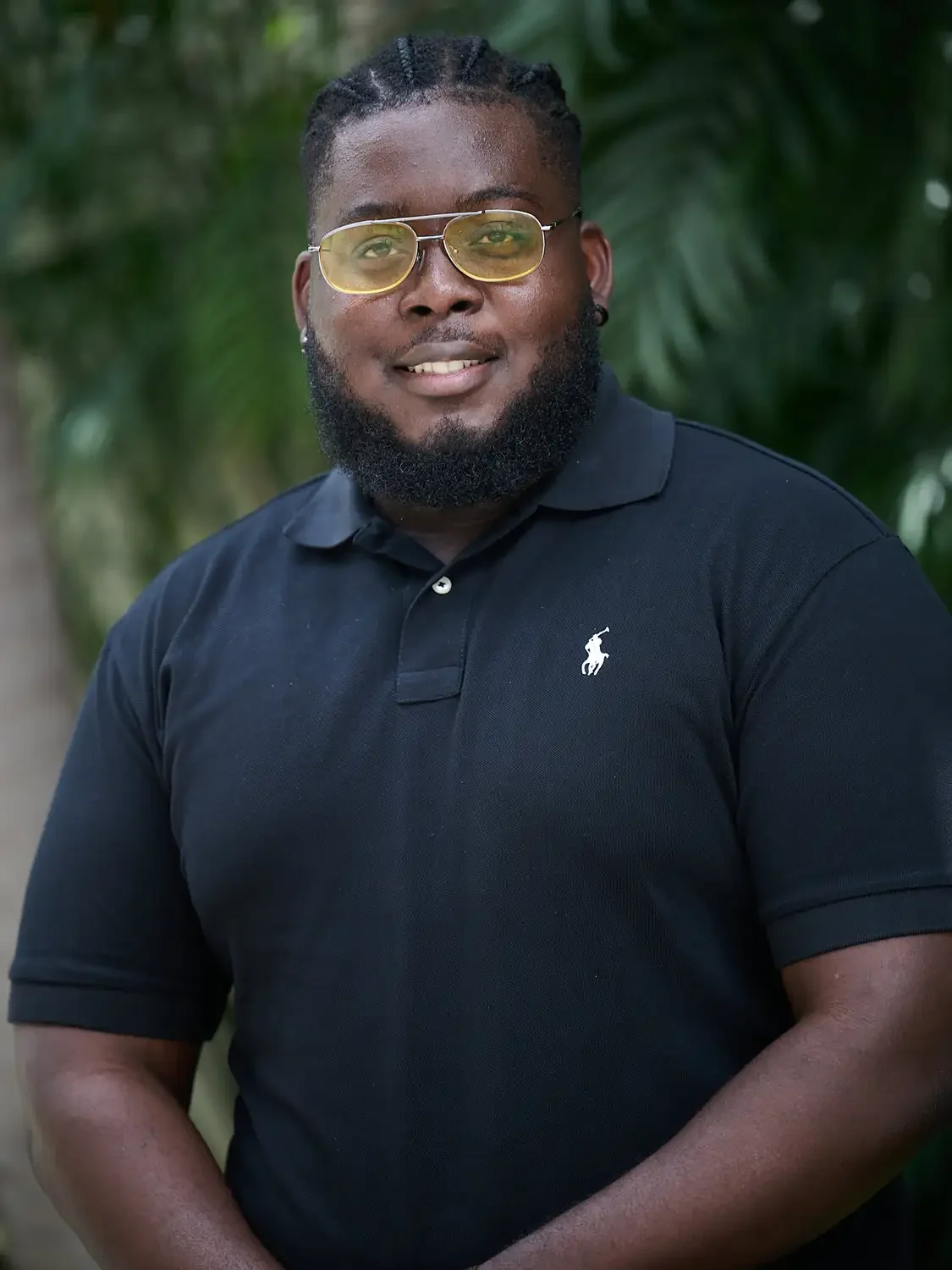 A man with glasses and a beard wearing a black polo shirt with a Ralph Lauren logo, standing outdoors Photographed in a professional way with green foliage in the background.