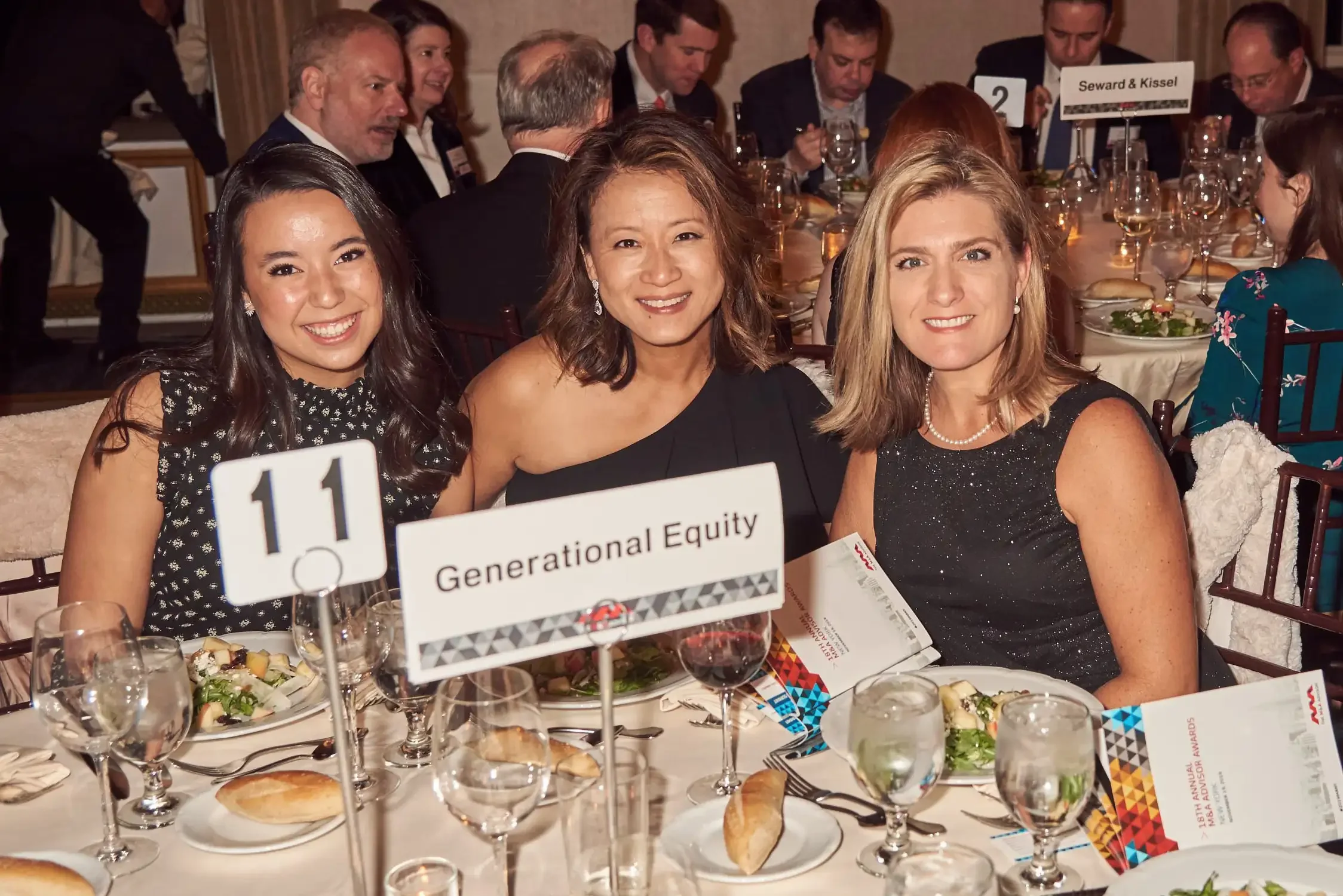People sitting at a corporate dinner table with food and drinks, smiling at the camera, with signs indicating different groups or categories.