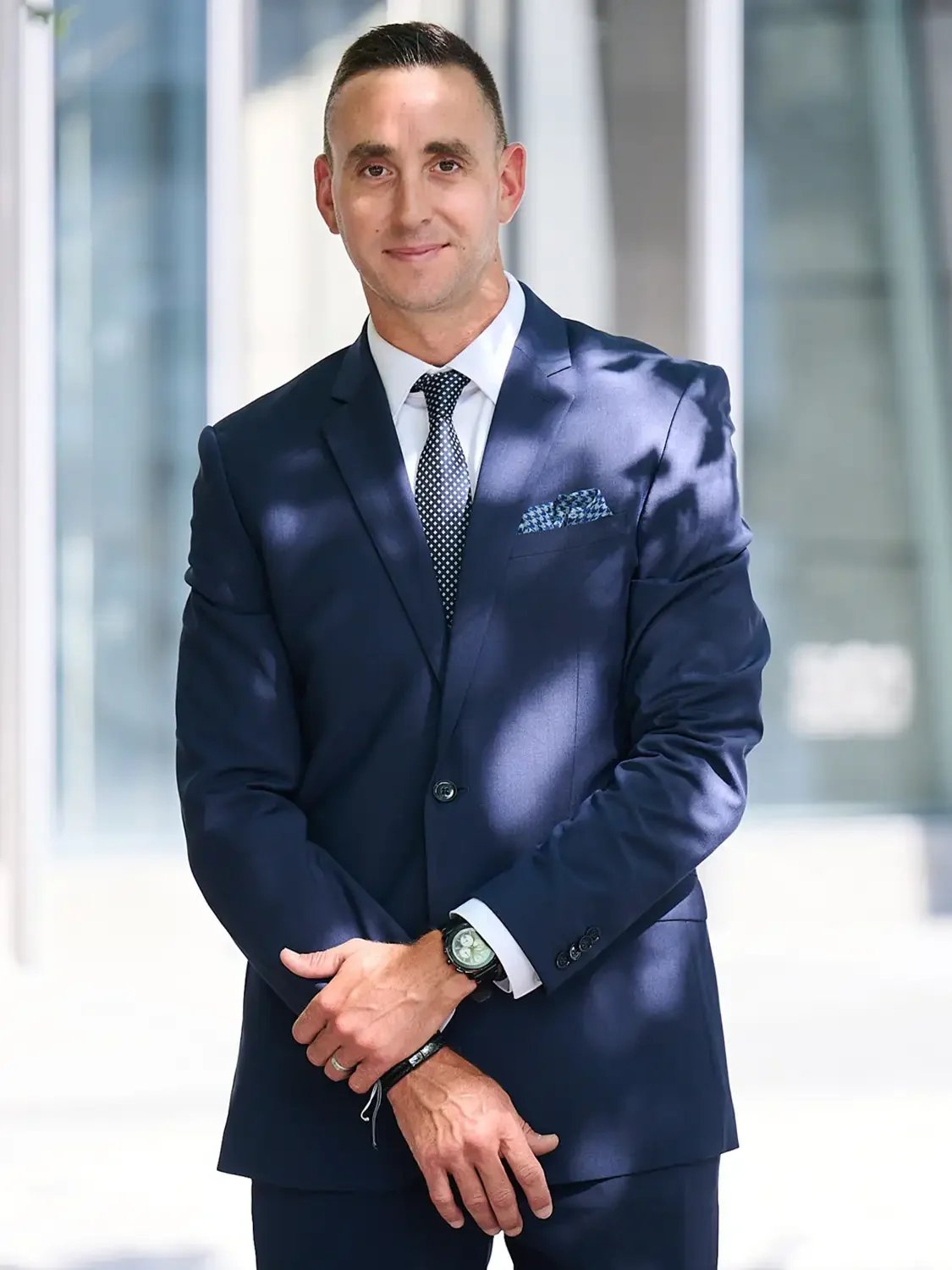 Outdoors headshot of a man in a navy blue suit standing in a modern building with glass walls.