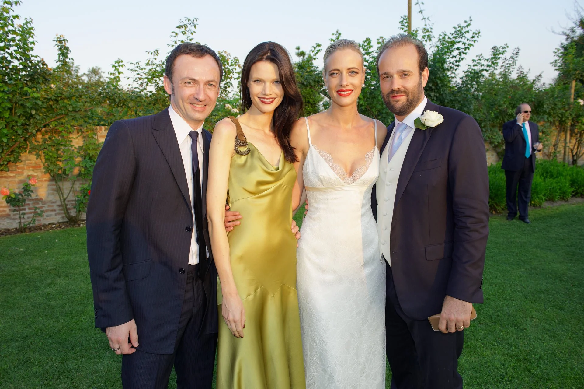 Four people dressed formally at an outdoor event, likely a wedding. Two men in dark suits and ties, two women in elegant dresses, one in a champagne-colored dress and the other in a white lace dress. The background features greenery and a man on a ph