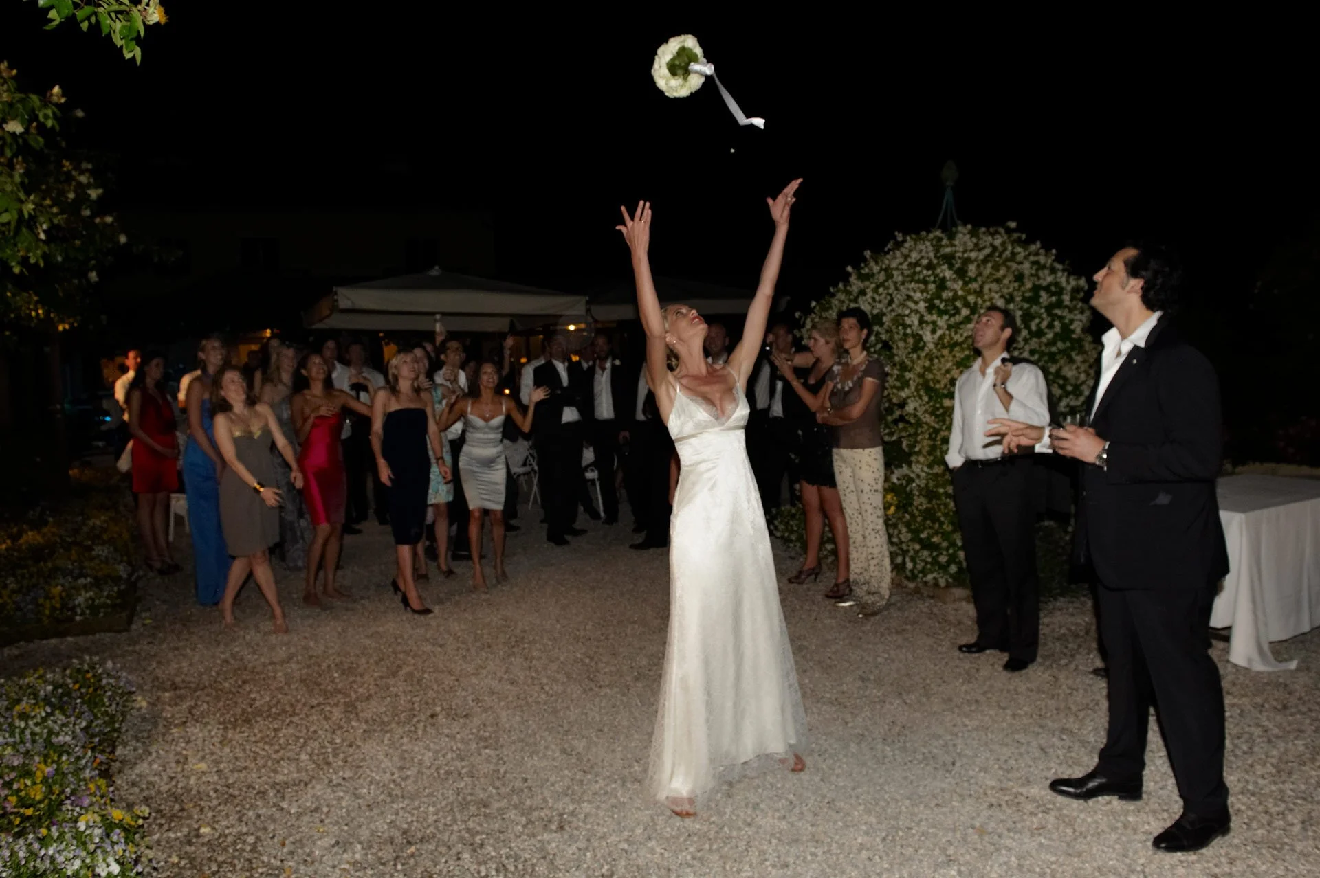 A bride wearing a white dress is throwing her bouquet into the air at a nighttime outdoor wedding reception, surrounded by guests.
