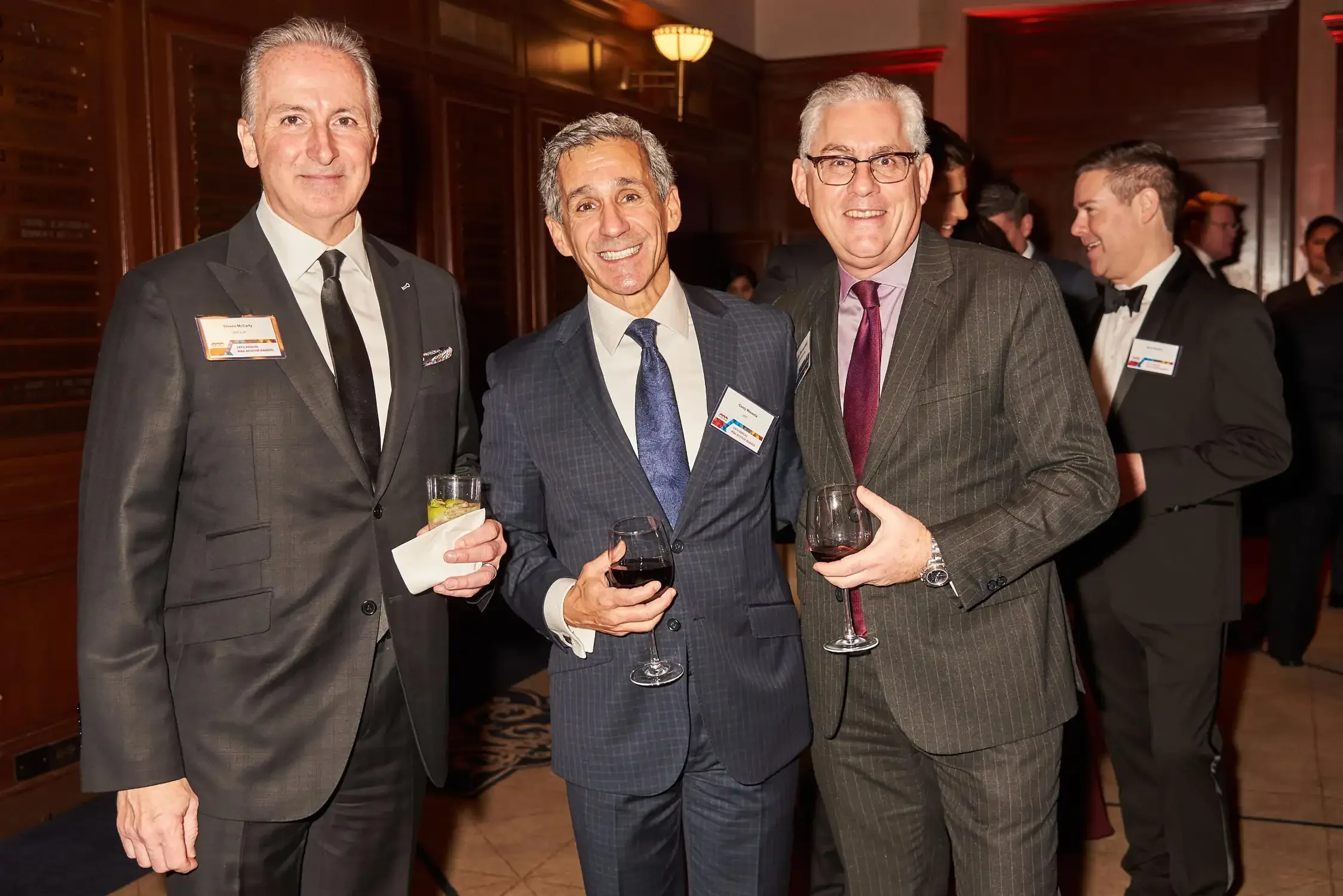 Three middle-aged men in suits smiling and holding glasses of wine at a formal corporate event, with other guests in the background.