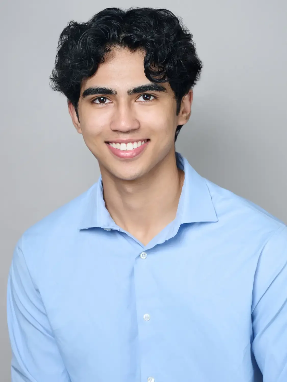Headshot of a  young man with black, curly hair, wearing a light blue button-up shirt, smiling against a studio lighting.