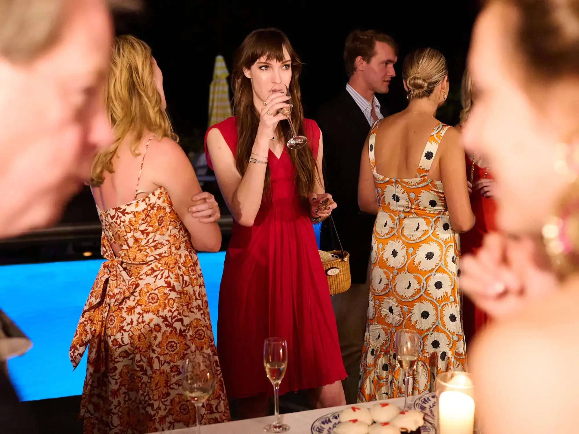 Group of people dressed in colorful dresses and suits gathered at a party, with wine glasses in hand and a table with a white tablecloth, candles, and food in the foreground.