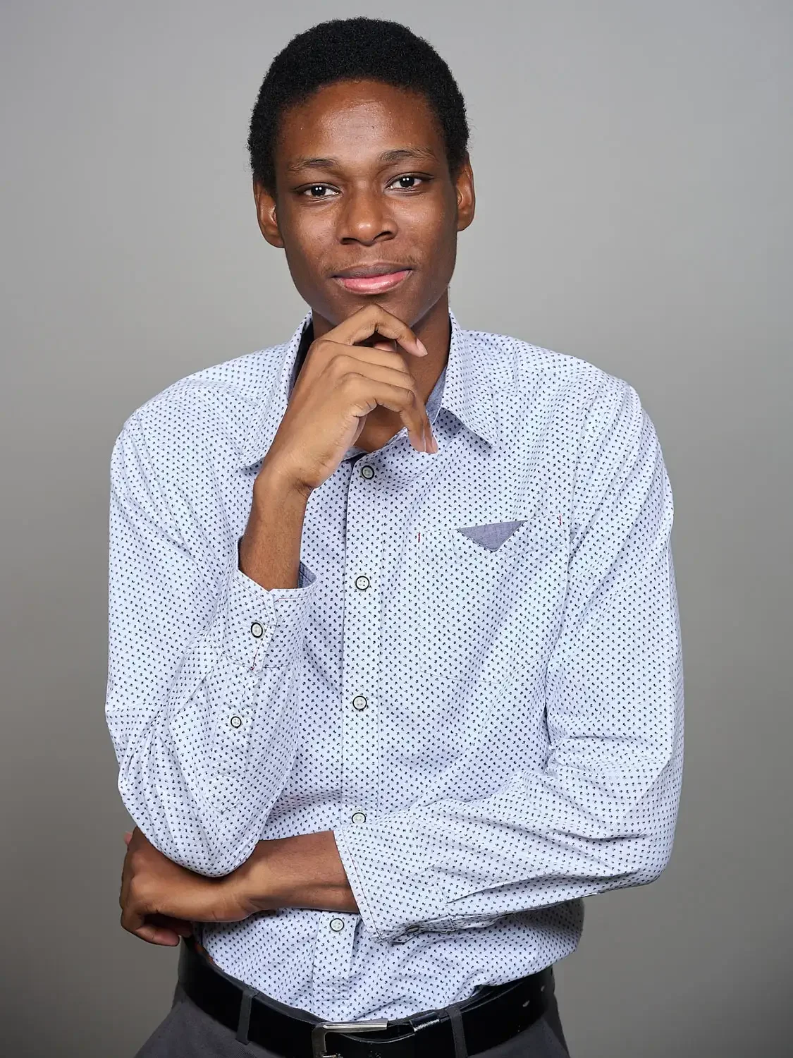 Headshot of a young man with short curly hair, wearing a white button-up shirt with small black dots, photographed in a professional photo studio. He is posing with friendly attitude and extreme confidence.