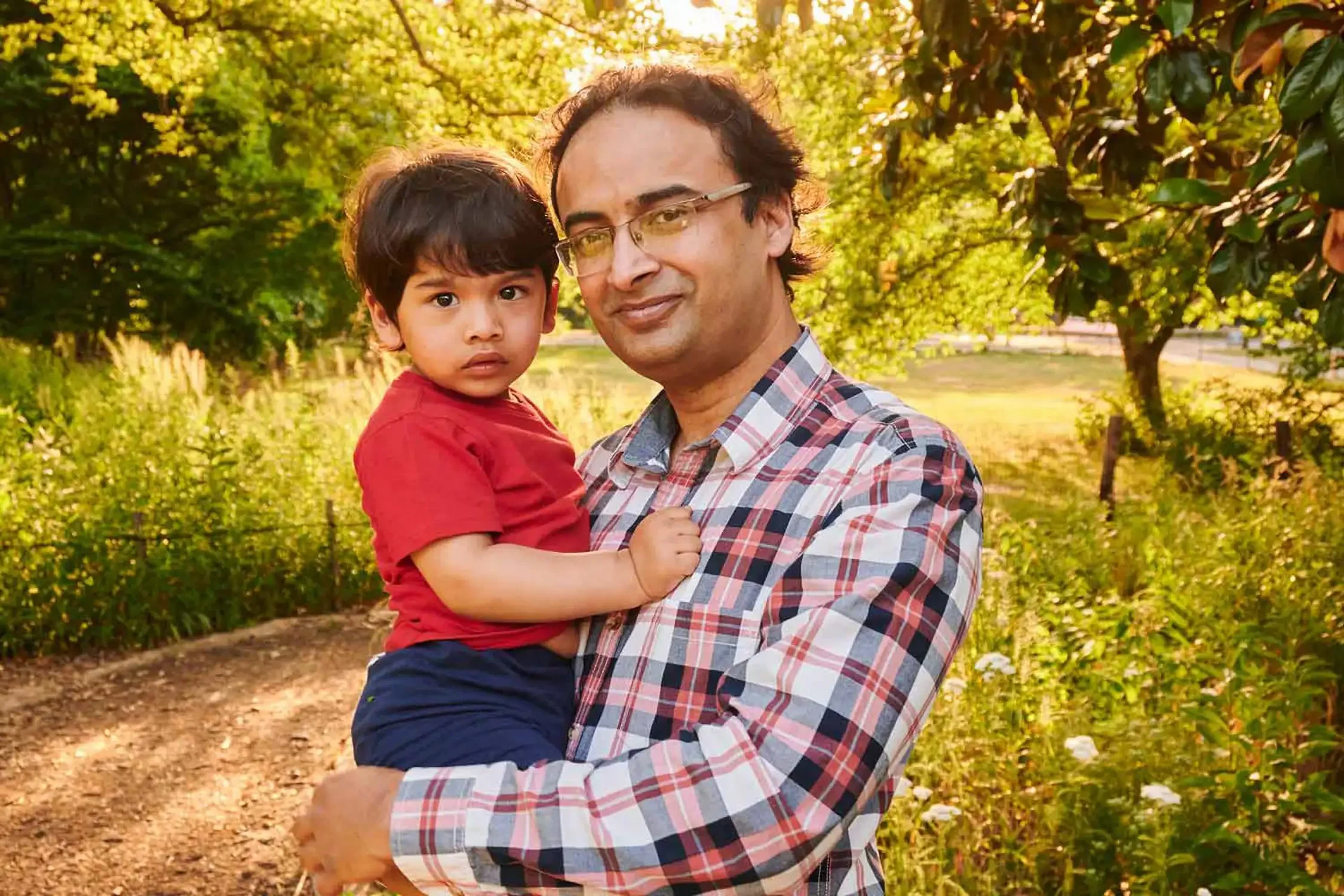 A man holding a young boy outdoors in a park during sunlight, with green trees and a dirt path in the background.