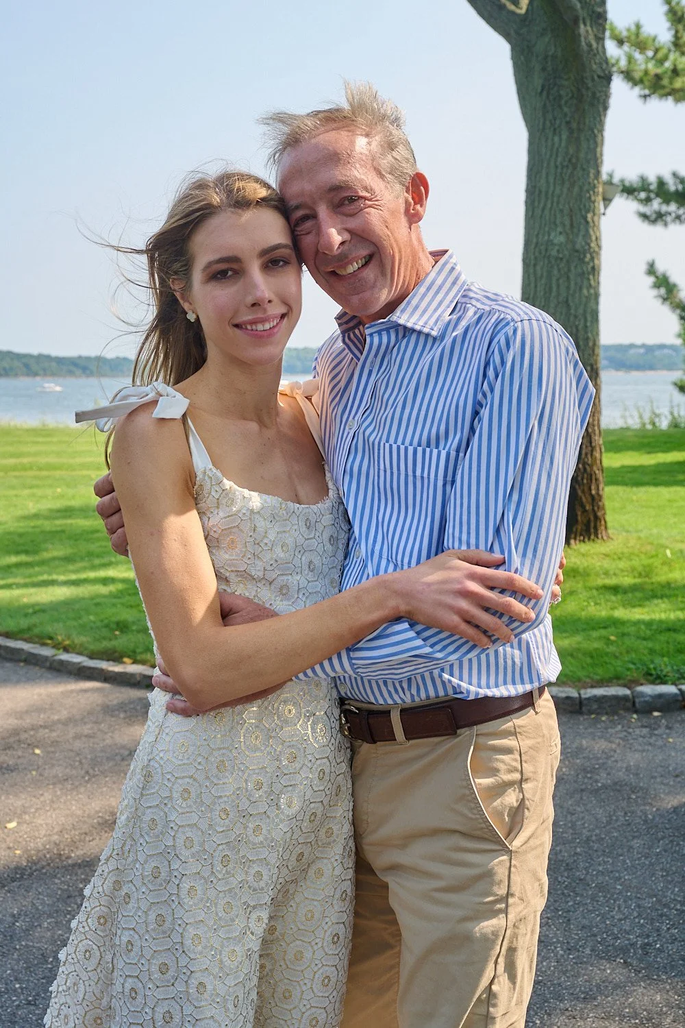 A young woman and an older man are standing close together outdoors, smiling, with trees and a body of water in the background.