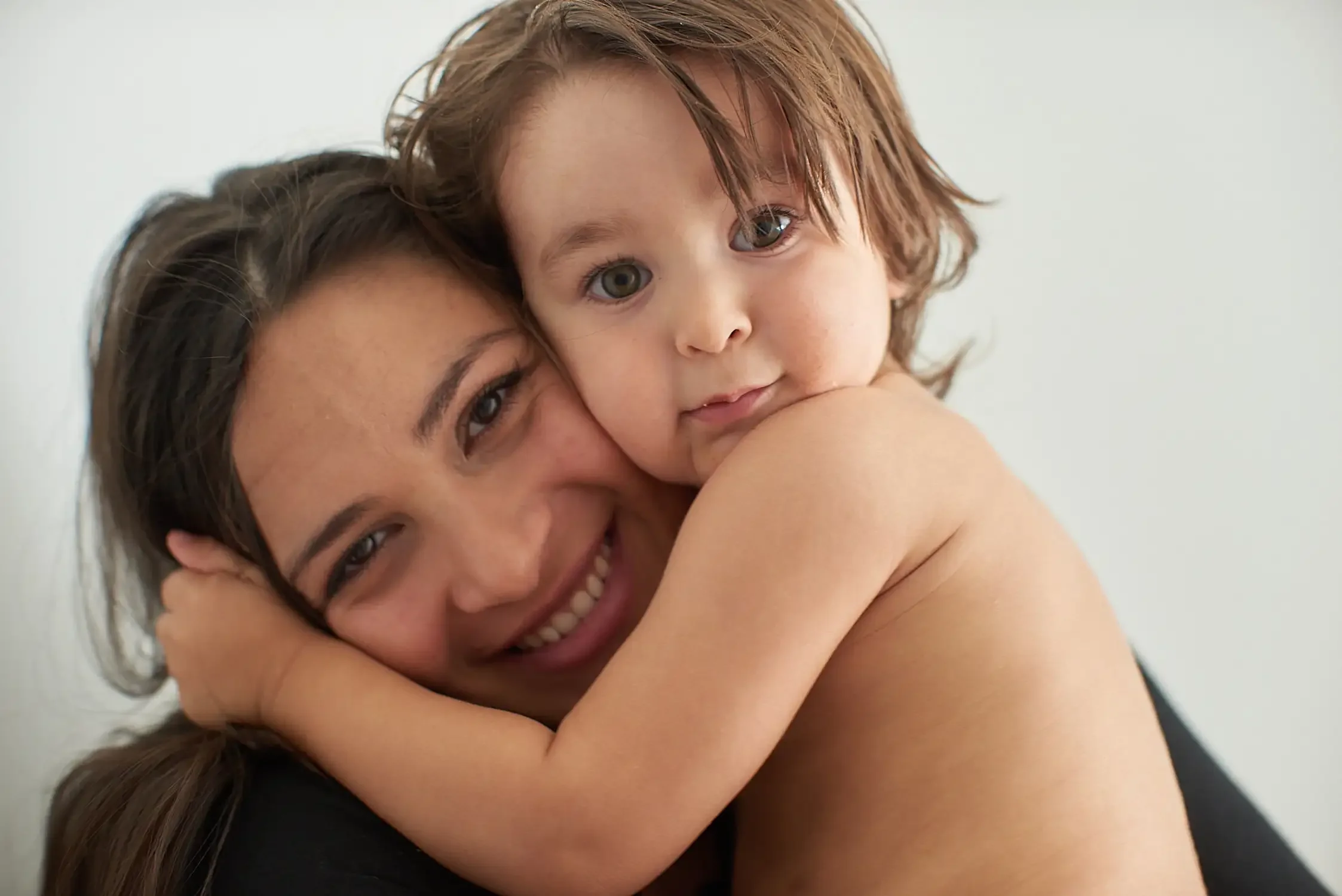 A woman with long dark hair and a big smile hugging her young boy with red hair and gray eyes, both looking closely at the camera in their home.