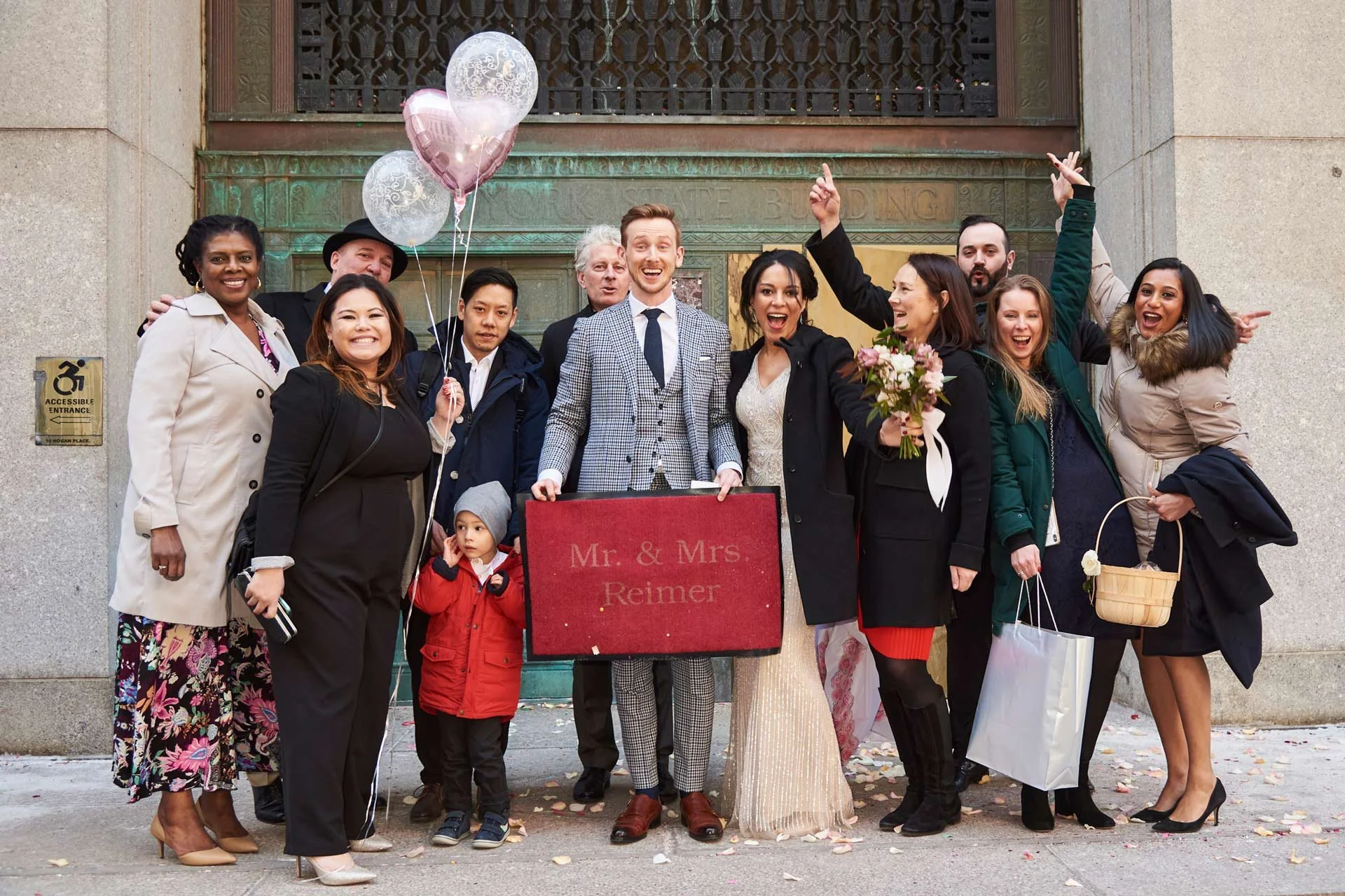 A group of people, including the bride and groom, celebrating in front of a building after a wedding. They are smiling, holding balloons, flowers, and gift bags, with confetti on the ground.