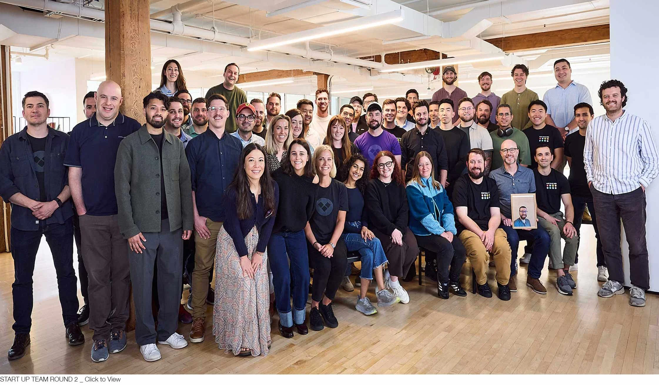 Large group of diverse young adults smiling for a team photo in a modern office space with wooden beams and light-colored walls.