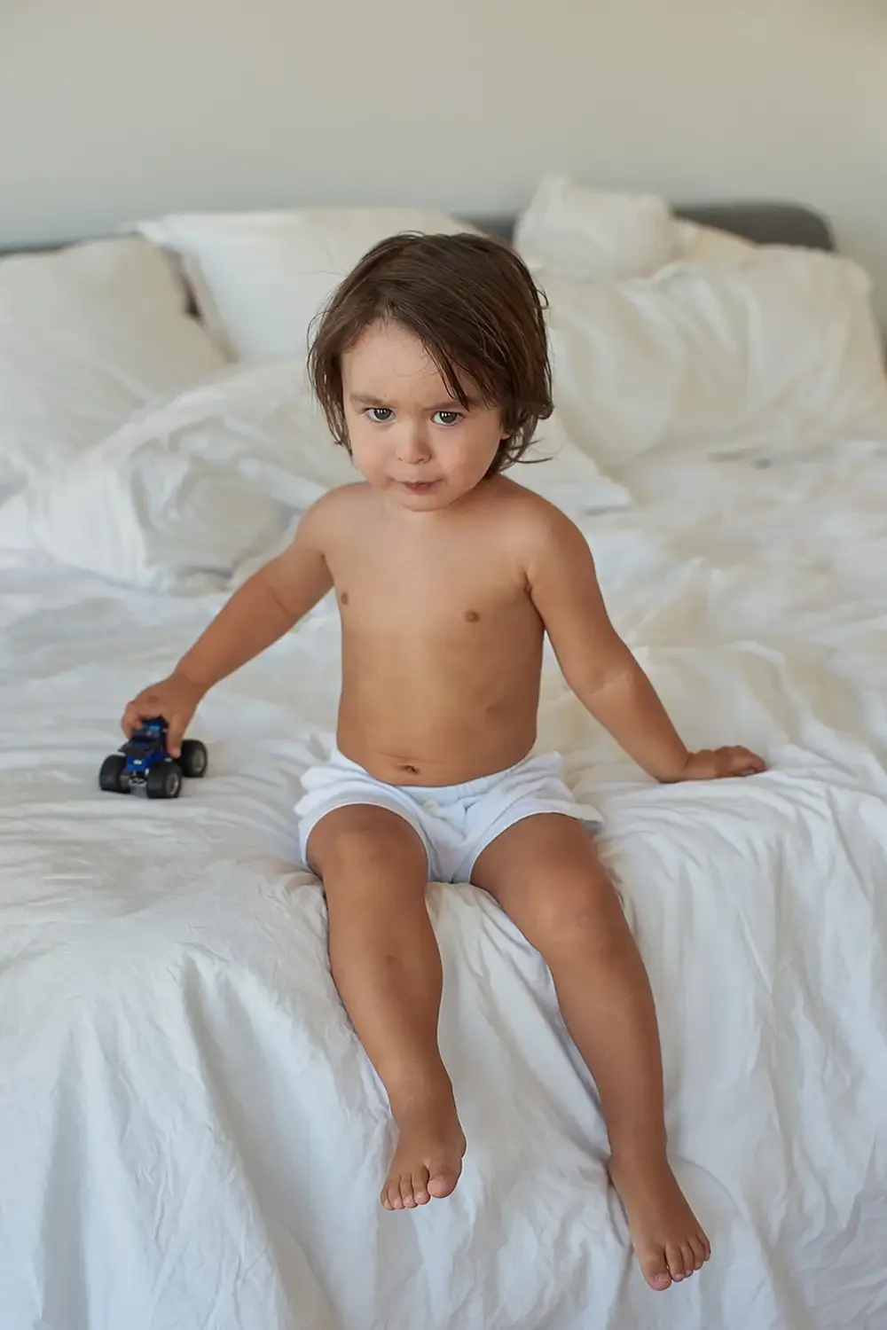 A young boy with brown hair and blue eyes sitting on a bed, holding a small blue toy car, wearing only white shorts, with a fun expression.