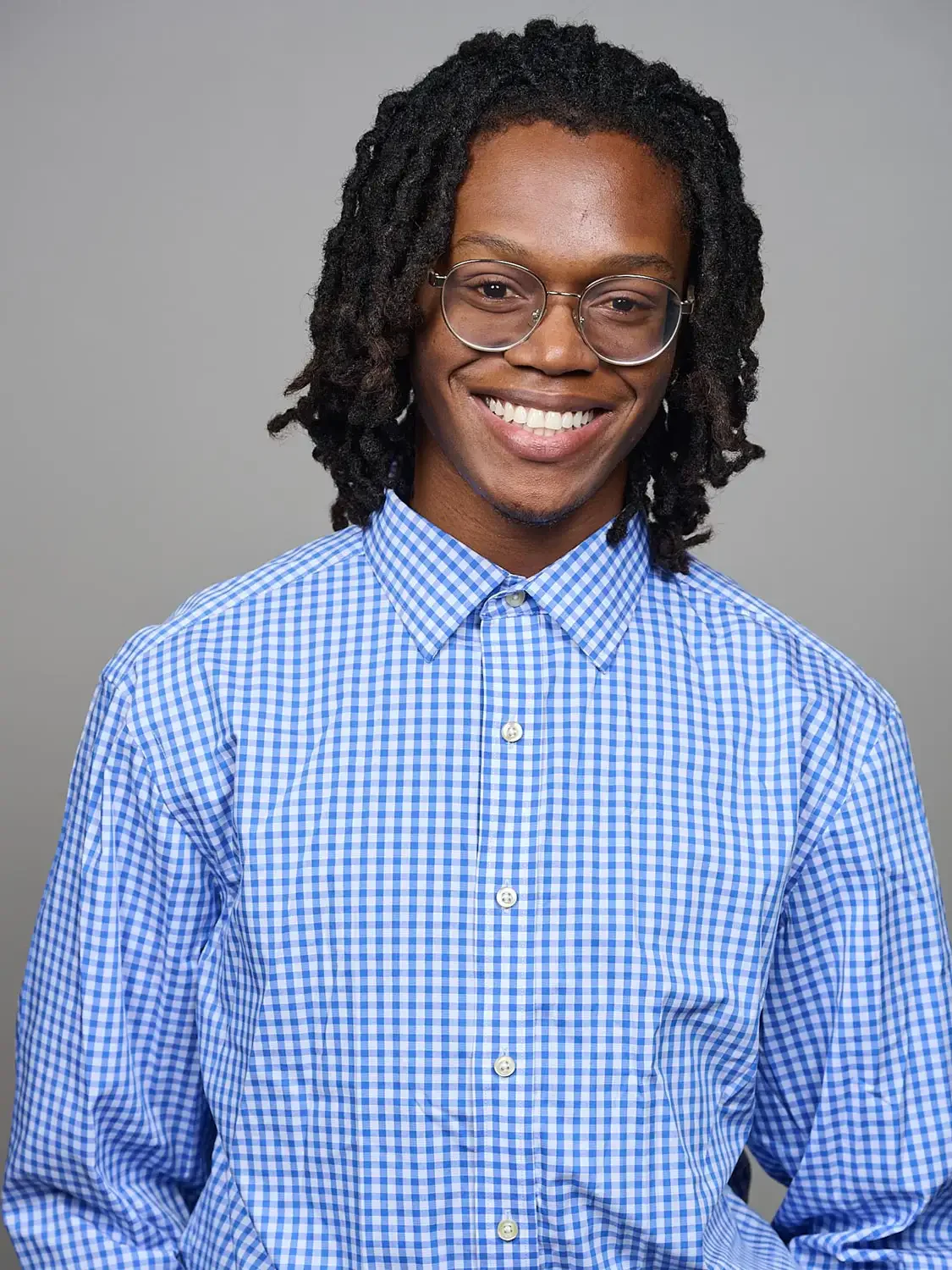 Portrait of a young man with dark curly hair, glasses, and a blue checkered shirt smiling in a photo-studio environment.