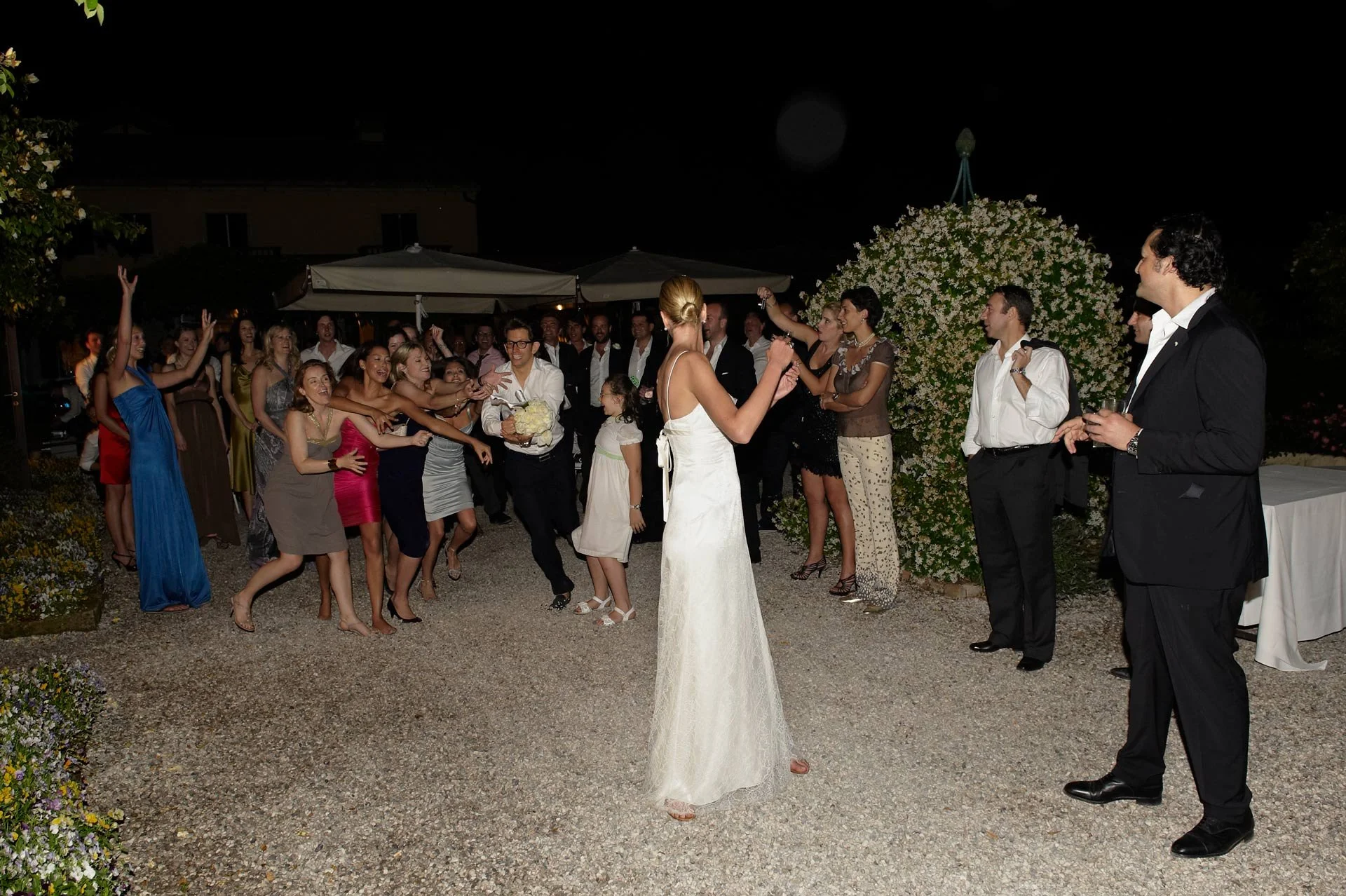 Bride and groom dancing at an outdoor wedding reception at night, surrounded by dancing guests and floral decorations.