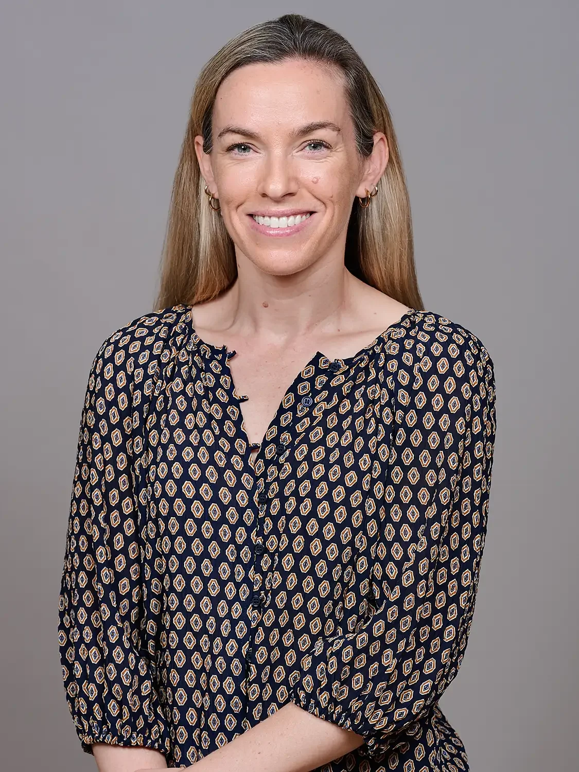 Headshot of a woman with long light brown hair and blue eyes smiling at the camera. She is wearing a dark patterned blouse with puffy sleeves and a v-neck.