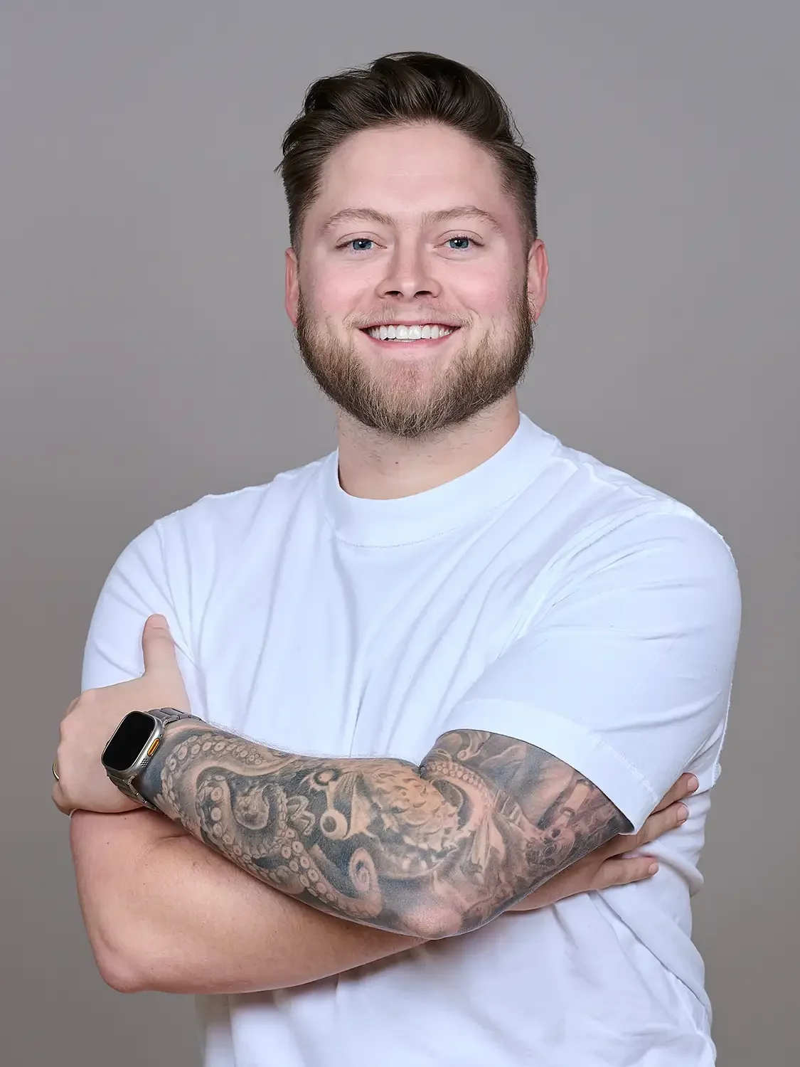 Headshot of a  smiling young man with a beard and short, styled hair, wearing a white T-shirt, is crossing his arms. He has detailed tattoos on his left arm smiling confident.
