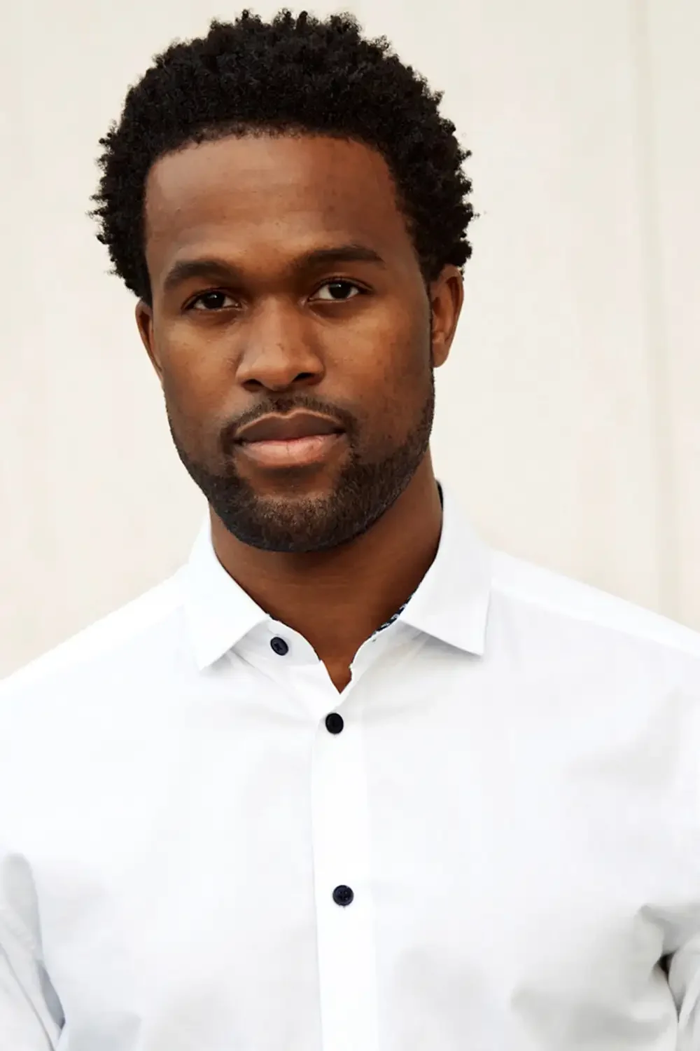 Portrait of a Black man with curly hair, wearing a white collared shirt, in an outdoor setup. Friendly