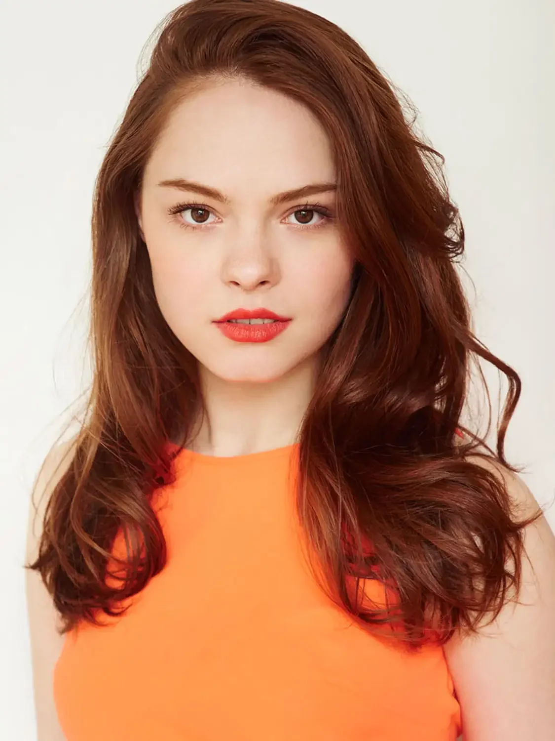 A young woman with wavy red hair, fair skin, wearing orange clothing, photographed in a. professional photo studio
