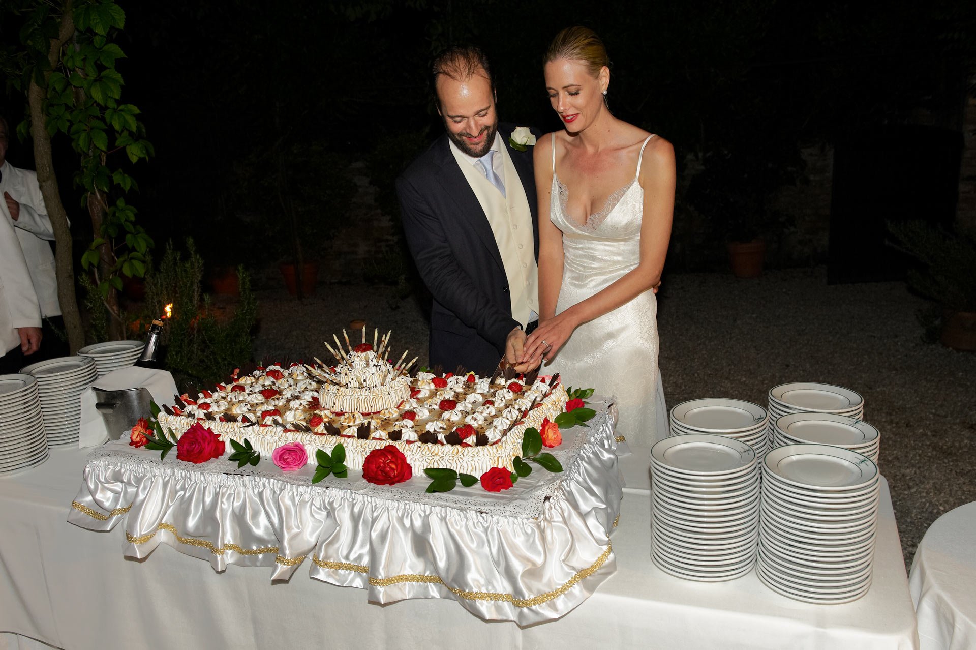 A bride and groom cutting a wedding cake together at a nighttime outdoor reception. The cake is decorated with flowers and a central cake topper, surrounded by plates and additional desserts.
