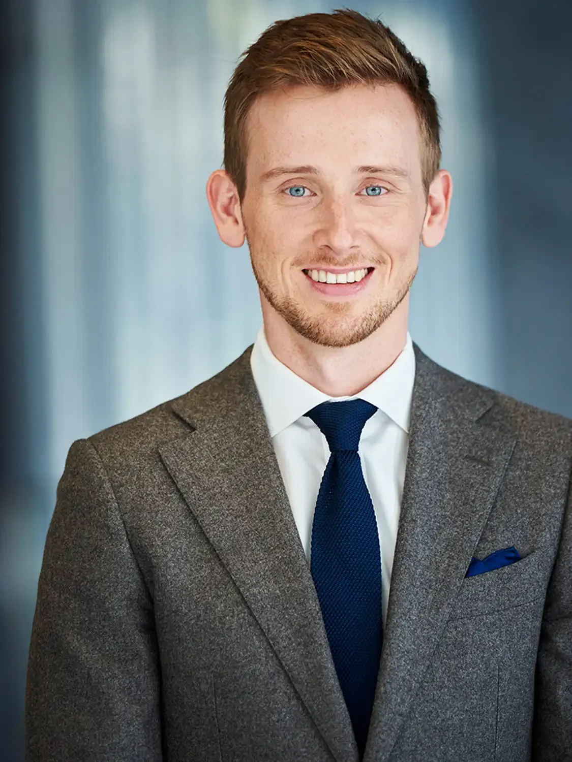 Portrait of a young man with light brown hair, blue eyes, and a beard, wearing a gray suit, white dress shirt, and a navy blue tie, smiling in a professional setting.