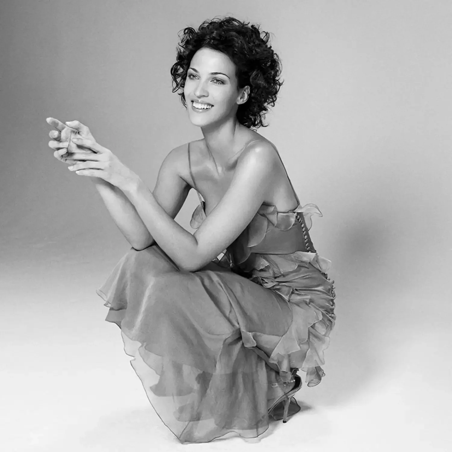Black and white photo shoot of a woman with short curly hair sitting on the floor, smiling, wearing a dress with ruffled skirt in a professional photo studio.