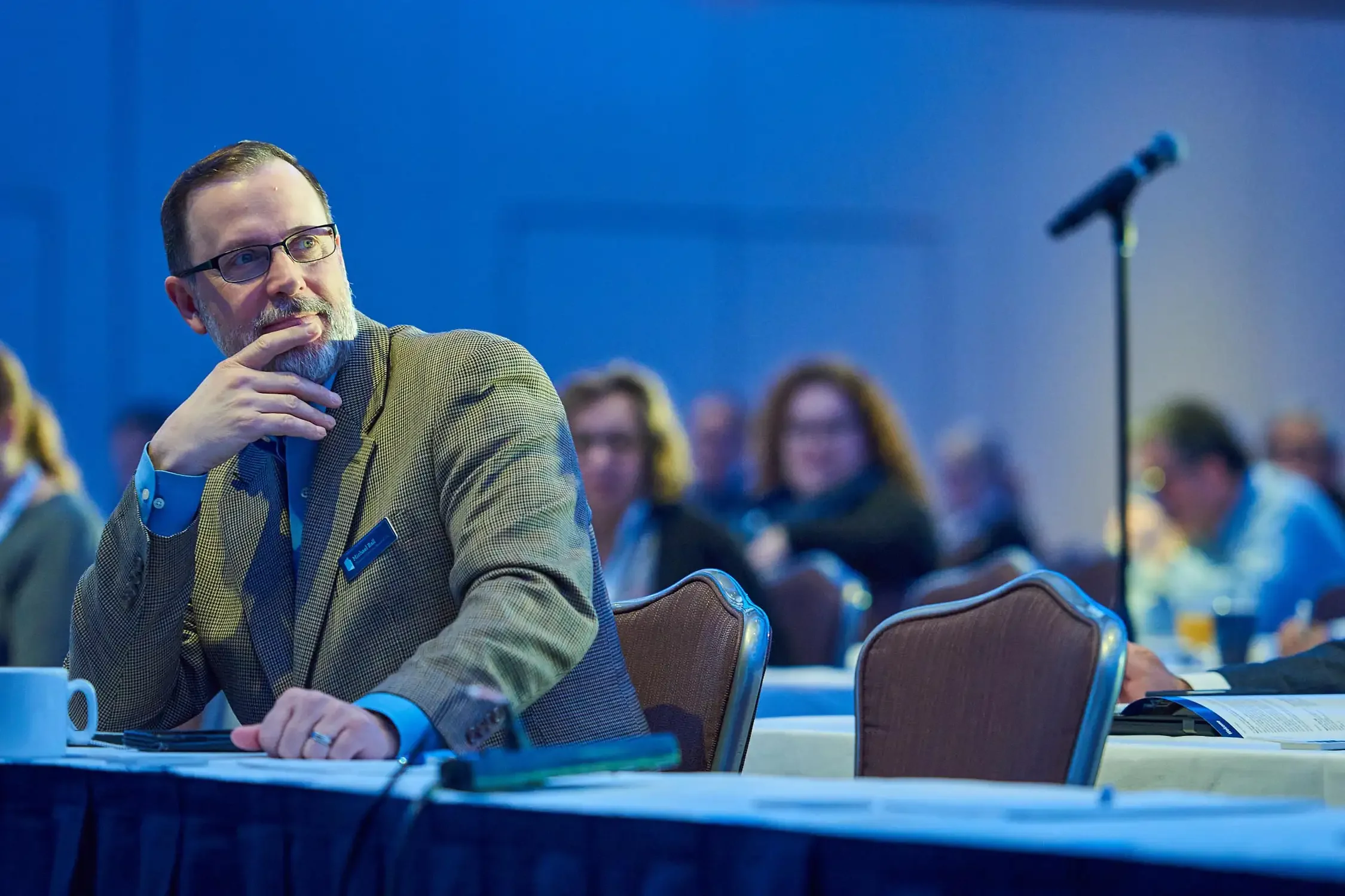 Corporate event photo of a man with glasses and a gray beard, wearing a brown blazer, sitting at a table during a conference. He is resting his chin on his hand.