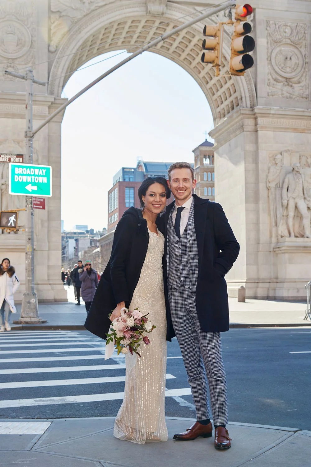 A smiling couple dressed in formal attire standing in front of an archway on a city street. The woman is holding a bouquet of flowers and is wearing a white dress with a black coat, while the man is wearing a checkered suit with a black coat. There a