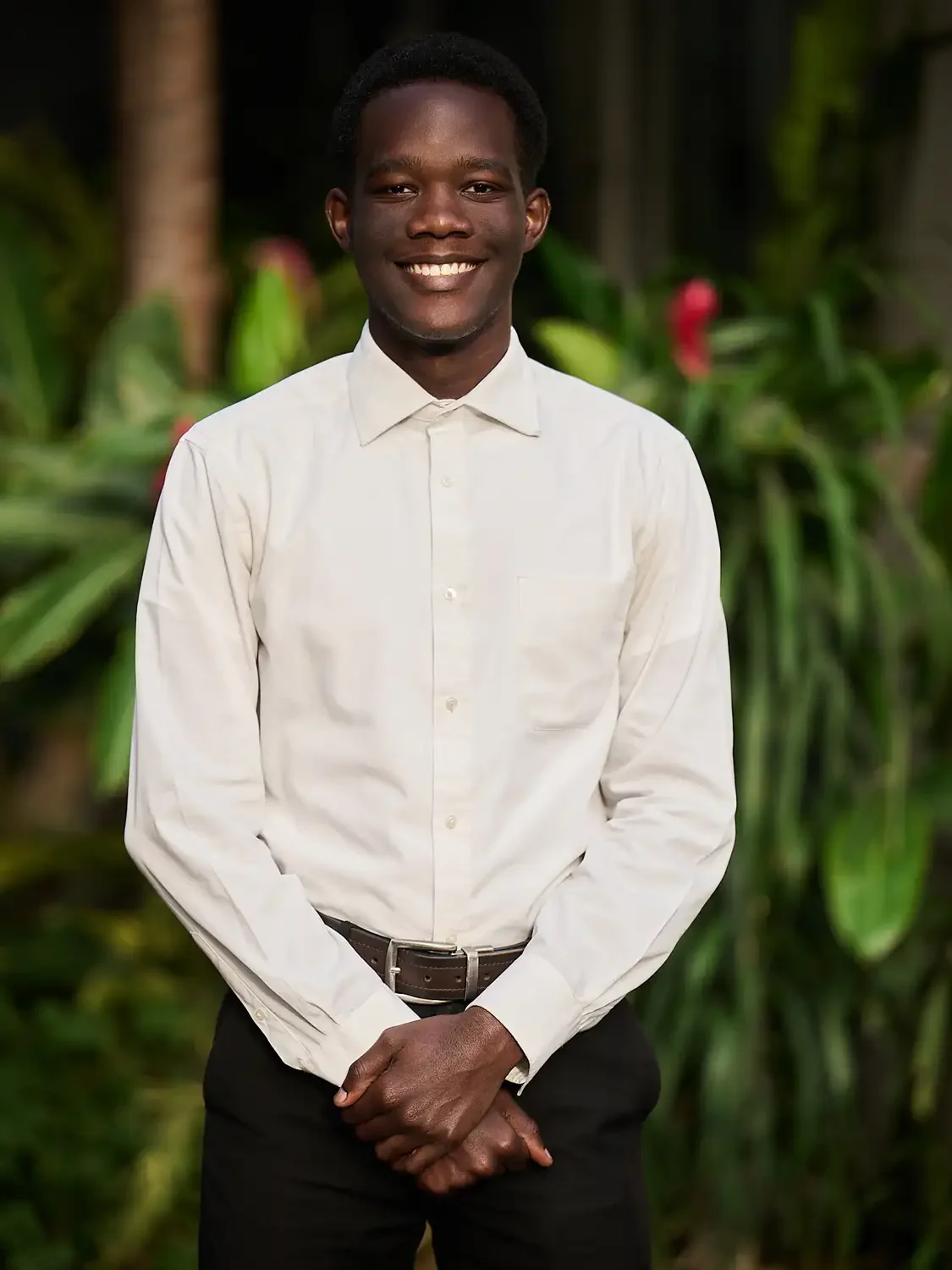 Portrait of a smiling man in a white dress shirt and black pants standing outdoors with green plants in the background.
