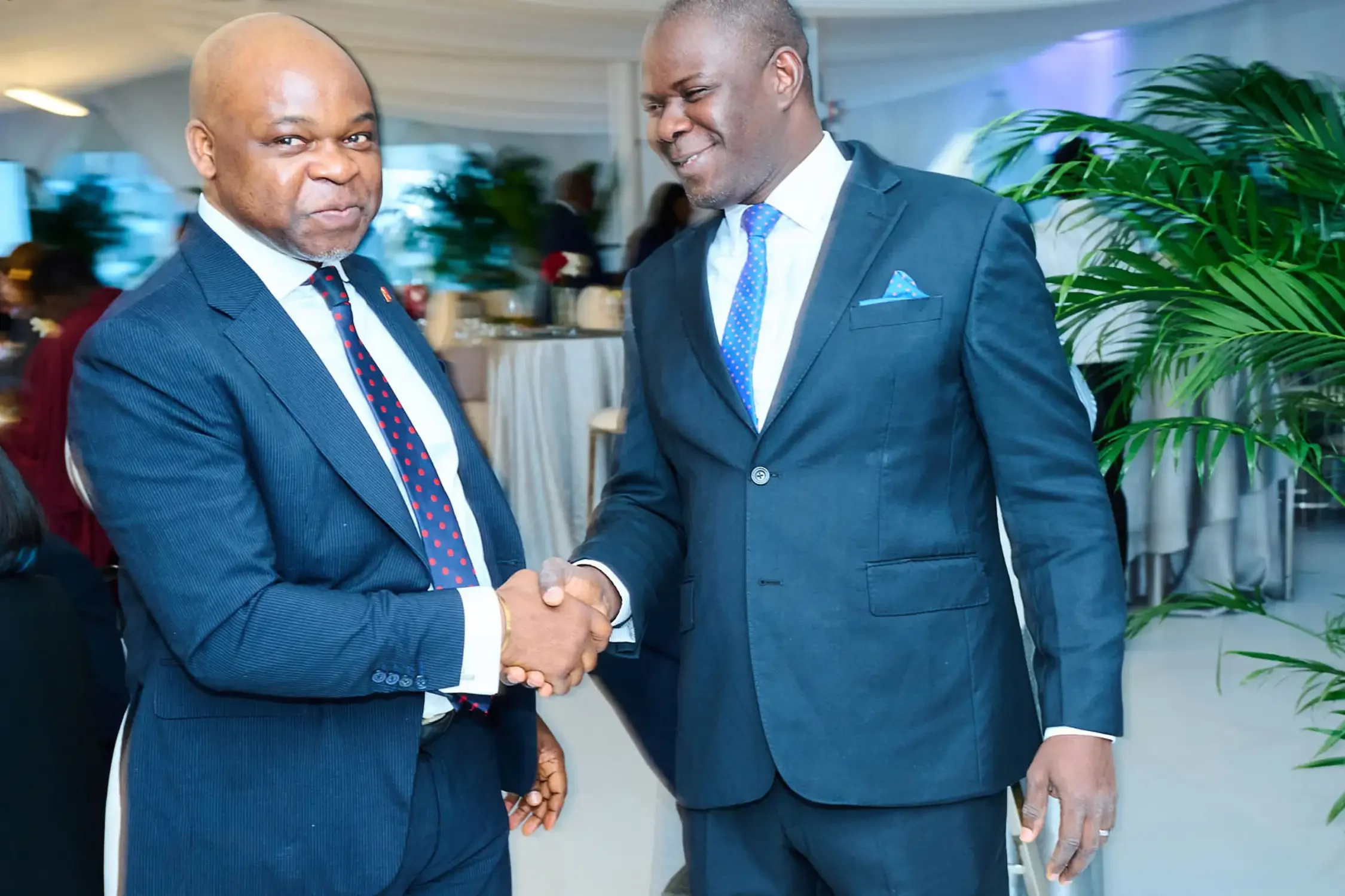 Two men in suits shaking hands at a formal event, smiling. One man is wearing a navy suit with a polka dot tie, and the other is wearing a dark gray suit with a blue polka dot tie and pocket square. Event photography