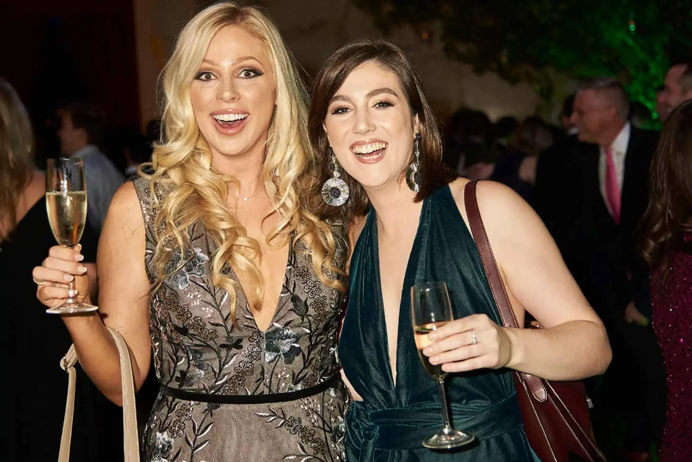 Two women smiling and holding glasses of champagne at a celebration or party, dressed in elegant attire with a crowd of people in the background.