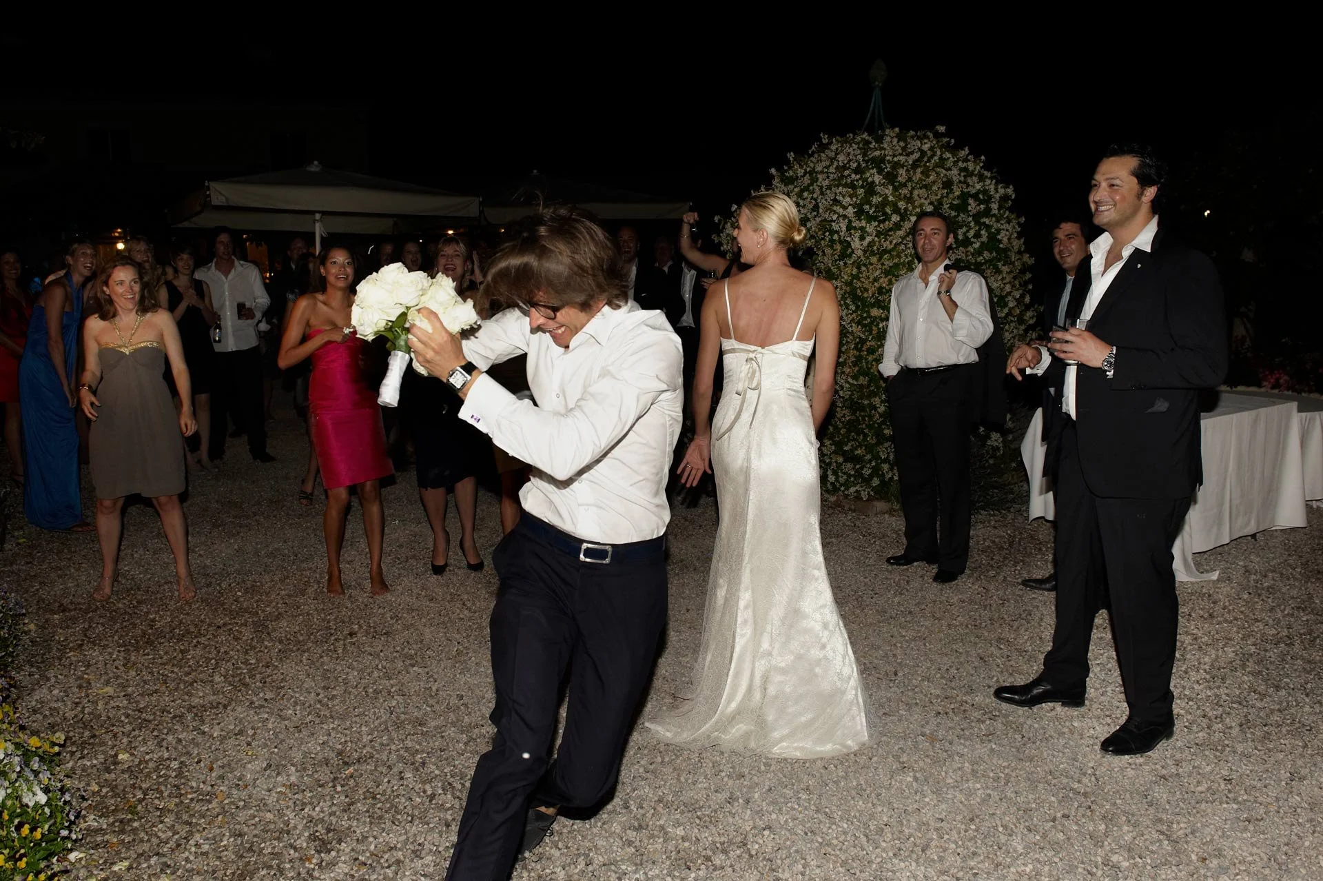 Guests celebrating at a wedding reception outdoors at night, with some women in dresses and men in suits, as a man dances and a woman in a white wedding gown holds a bouquet.