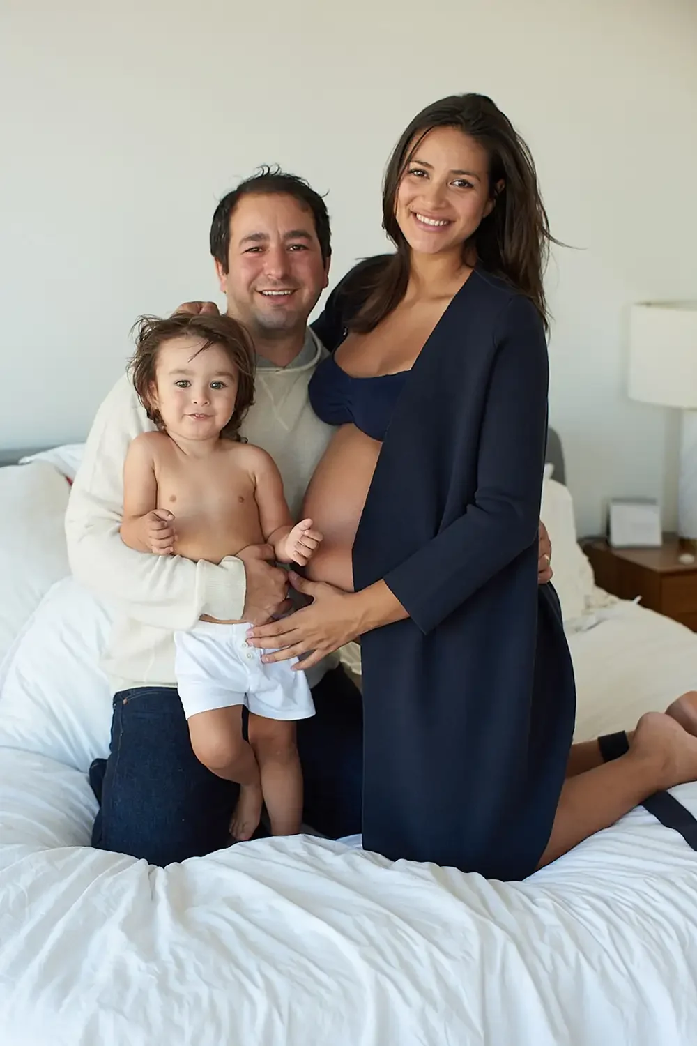 A happy family poses on a bed, with the mother showing her pregnant belly, the father holding their young child, all smiling at the camera in a cozy bedroom.