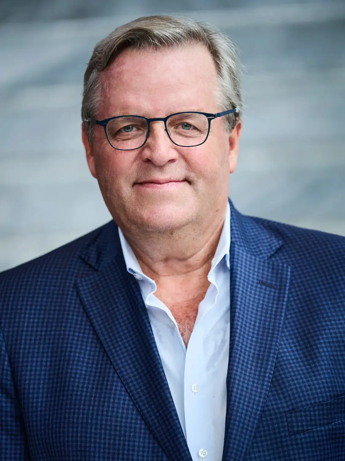 LinkedIn headshot of a startup founder in New York office of a middle-aged man with gray hair, wearing glasses smiling in front of a gray blurred background.