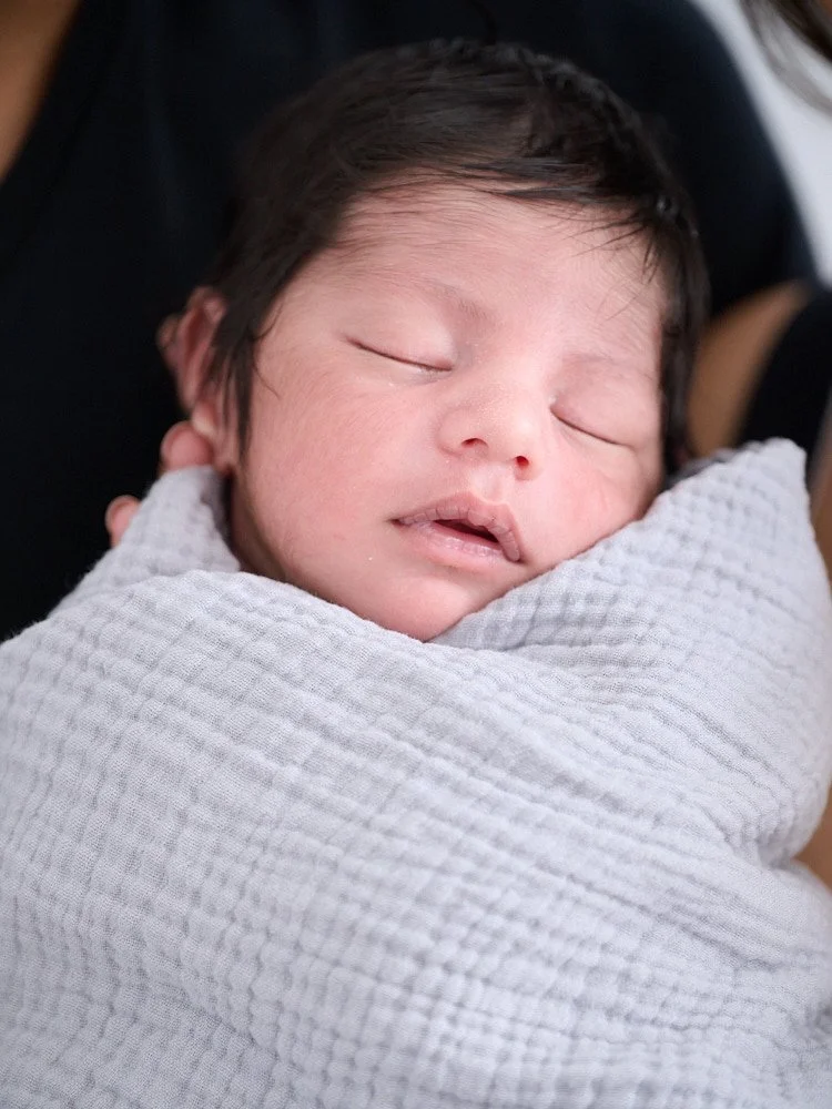 Close-up of a sleeping newborn baby with dark hair wrapped in a white blanket, being held by an adult.