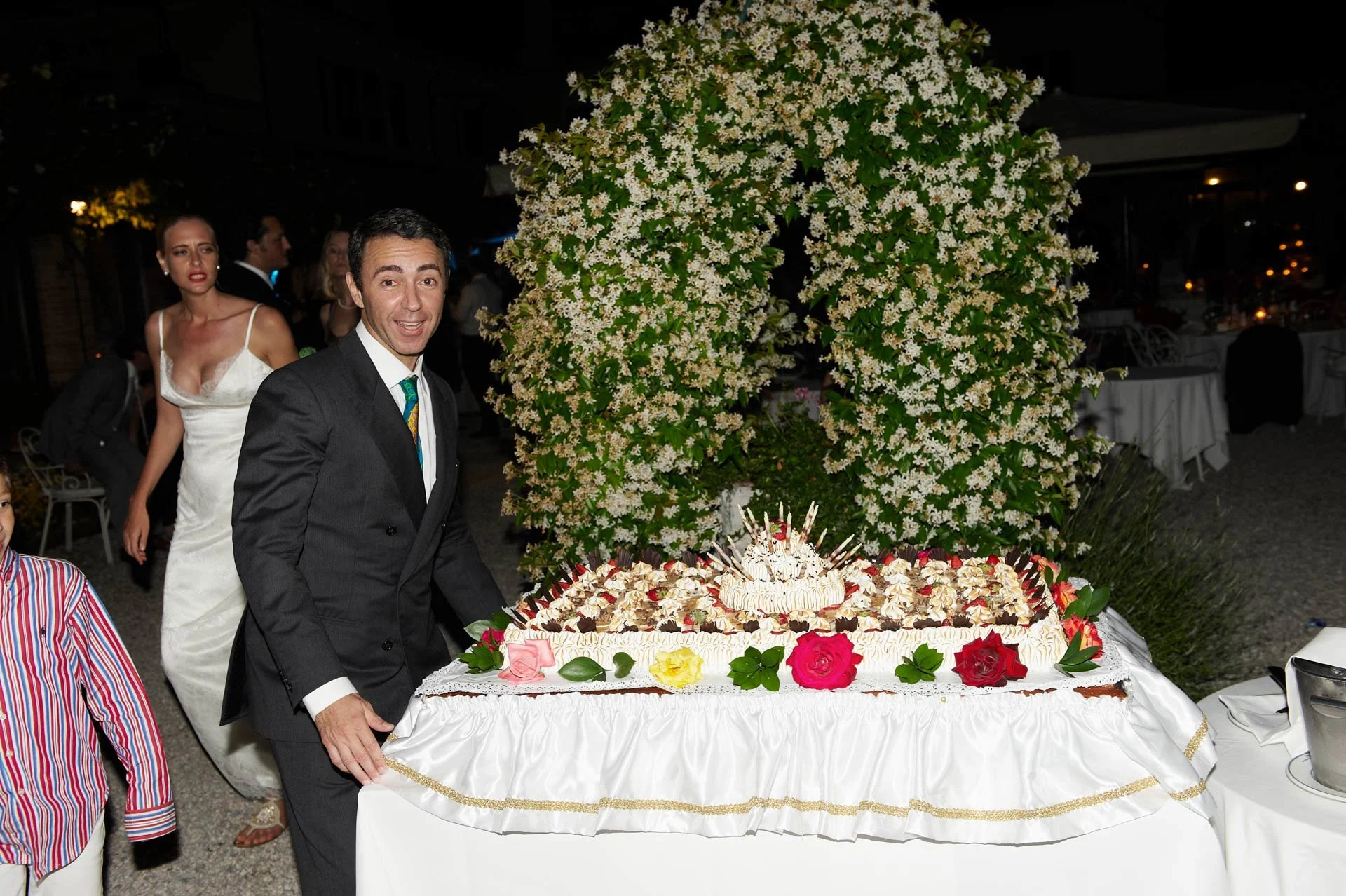 Man in a black suit and tie standing next to a large white wedding cake decorated with roses and other flowers, with a sizzling candle cake on top, at an outdoor wedding reception at night.