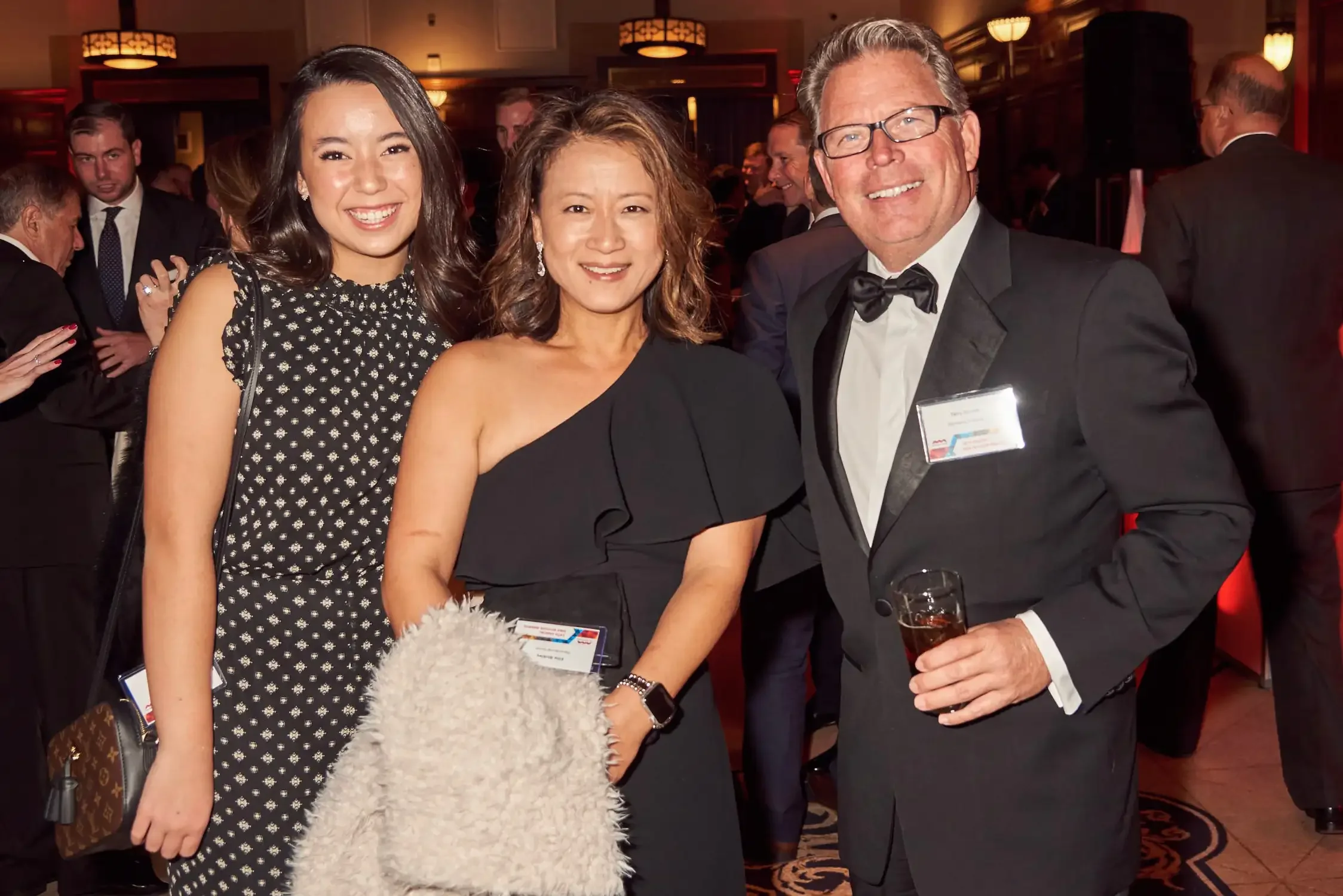 Three people at a formal event, two women and one man, posing and smiling for the camera; the man is holding a drink enjoying their time.