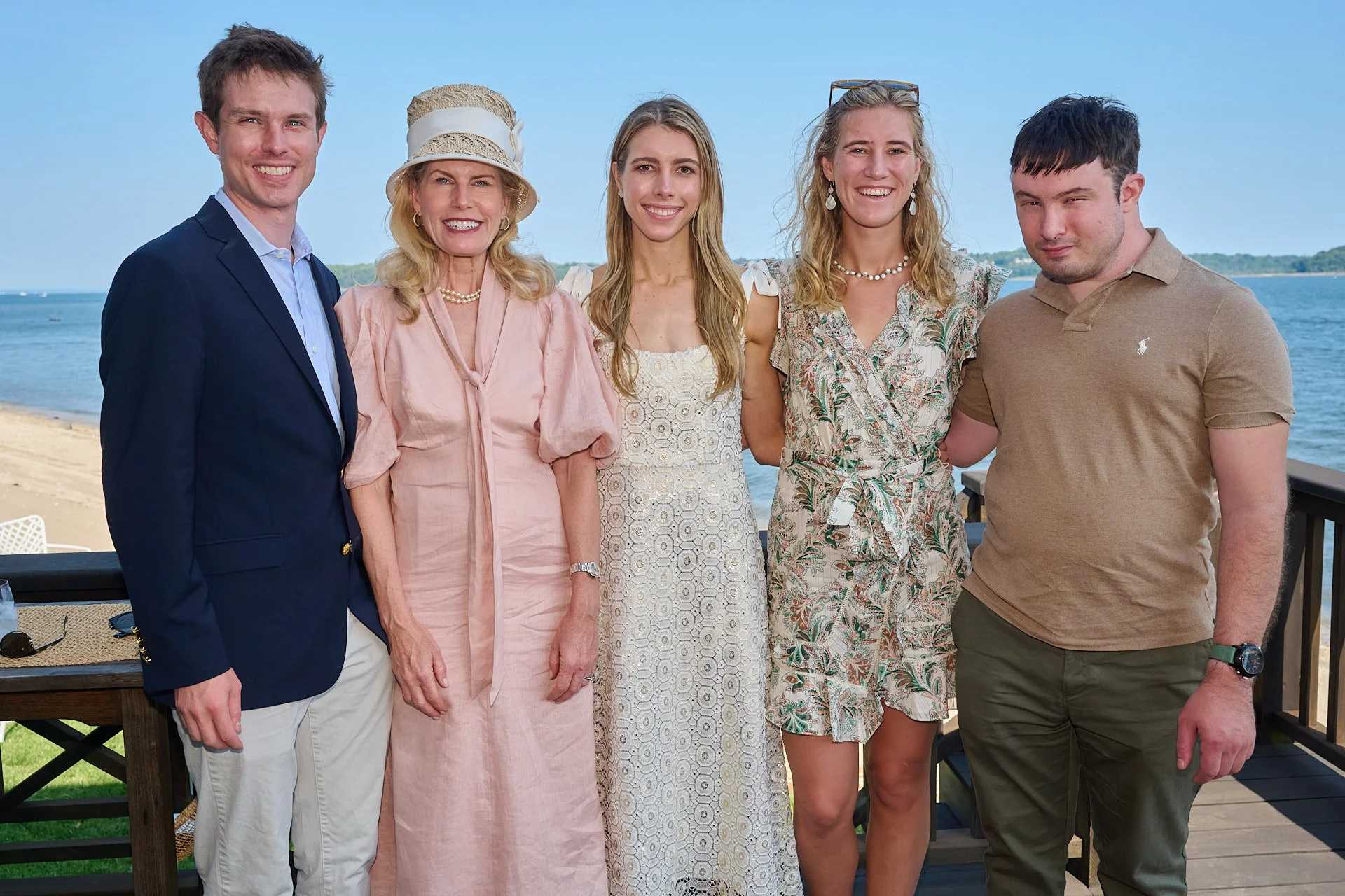 Group of five people standing close together on a beachside deck, smiling, with water and a distant shoreline in the background.