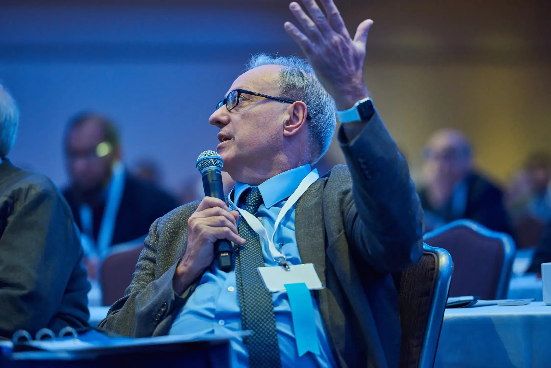 A man with gray hair, glasses, and a suit is speaking into a microphone at a conference. He is gesturing upward with his right hand and wearing a conference badge and smartwatch.