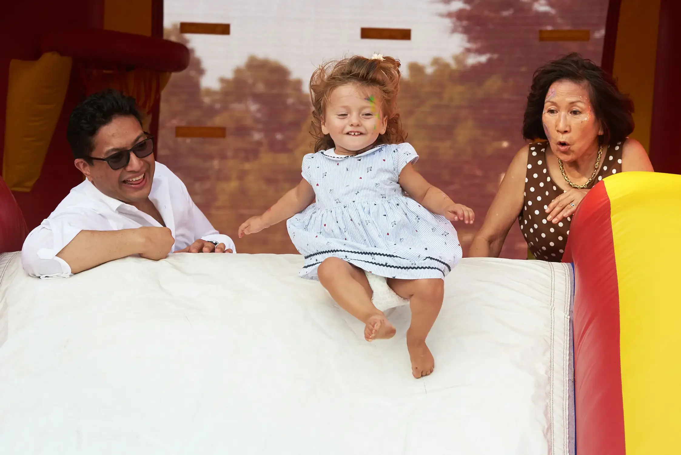 Family photographs of a young girl bouncing on a bed while two adults, a man and a woman, watch and smile. The girl is wearing a light blue dress with small designs.