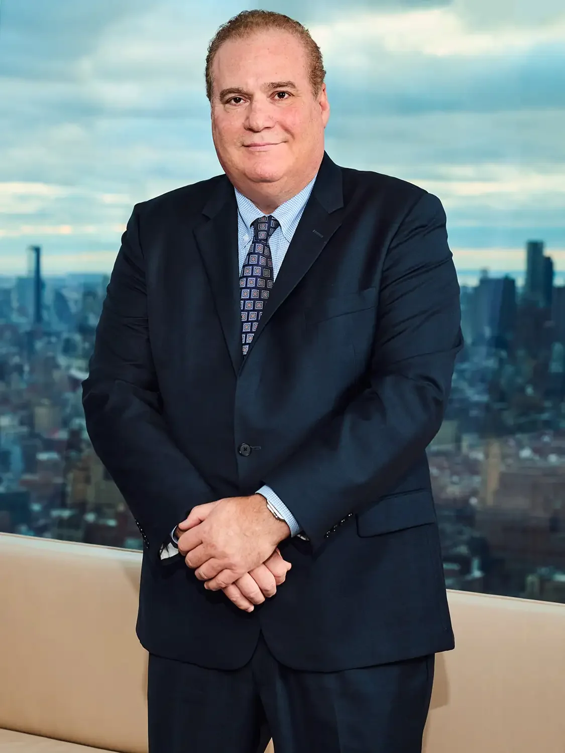 Portrait of a CEO in a dark suit with a patterned tie, standing indoors with a city skyline and cloudy sky in the background.